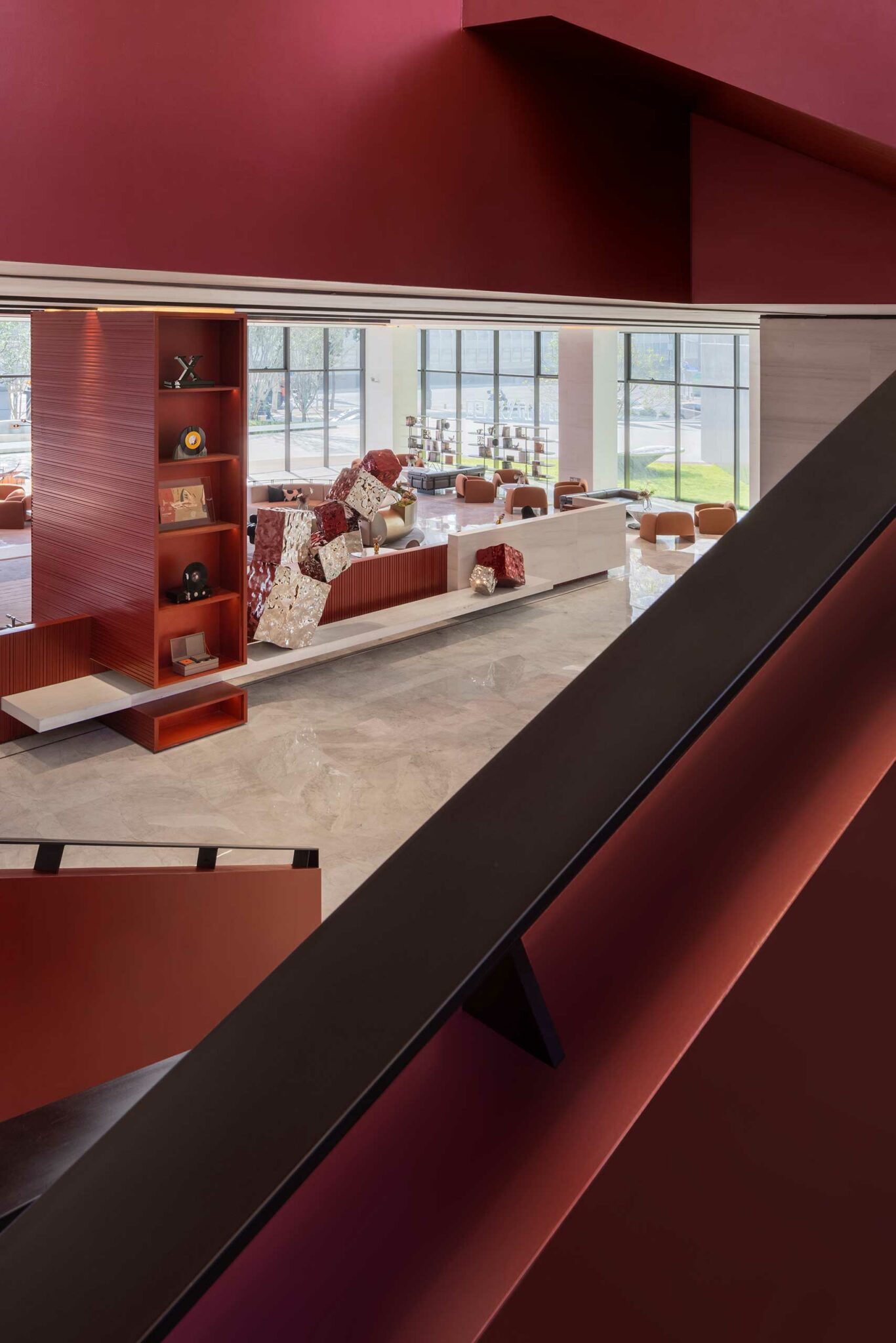 Modern interior with maroon and beige tones. Features geometric sculptures, open shelving, and floor-to-ceiling windows with a view of greenery.