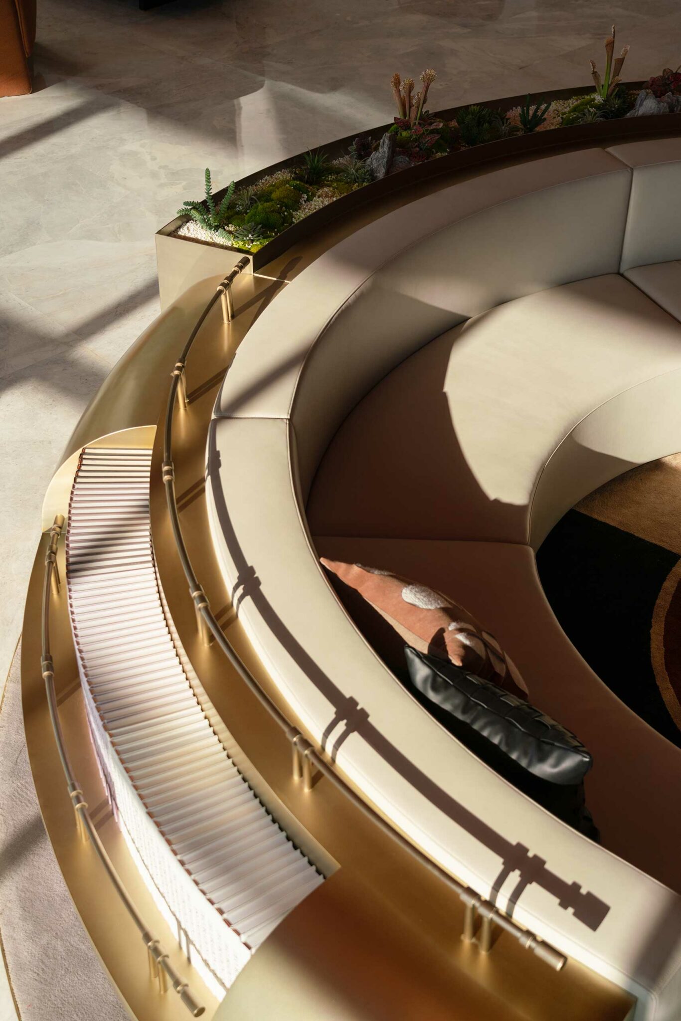 Curved beige seating with metal railings and throw pillows, next to a lush indoor planter with desert plants. Warm sunlight casts soft shadows.