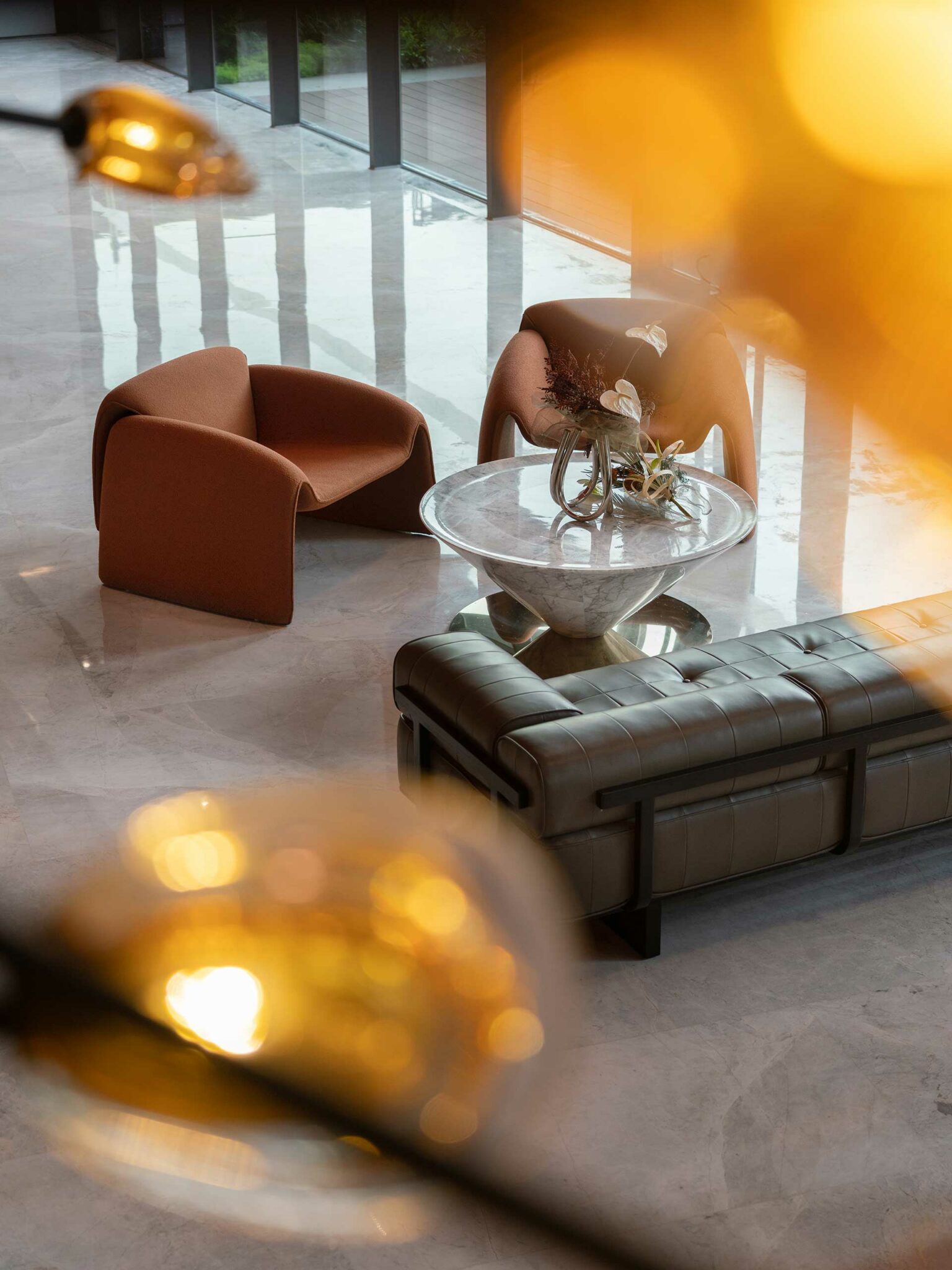 Modern lounge featuring a semi-circular white sofa, central marble and gold table with a glass vase, black chair, and earthy decor. Cozy and elegant.