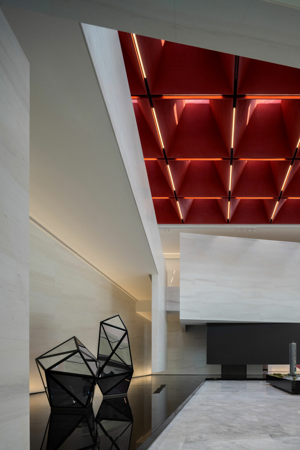 Modern interior with geometric design featuring a red grid ceiling, white walls, and two angular black sculptures on a glossy floor, creating a sleek, contemporary ambiance.