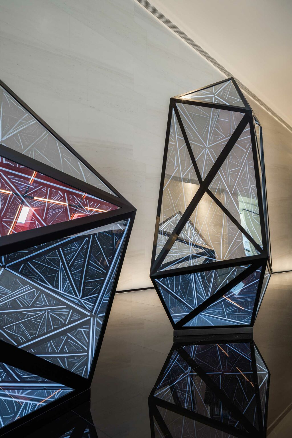 Geometric sculptures with mirrored surfaces and black frames reflect intricate patterns of light and color. The setting is modern and abstract.