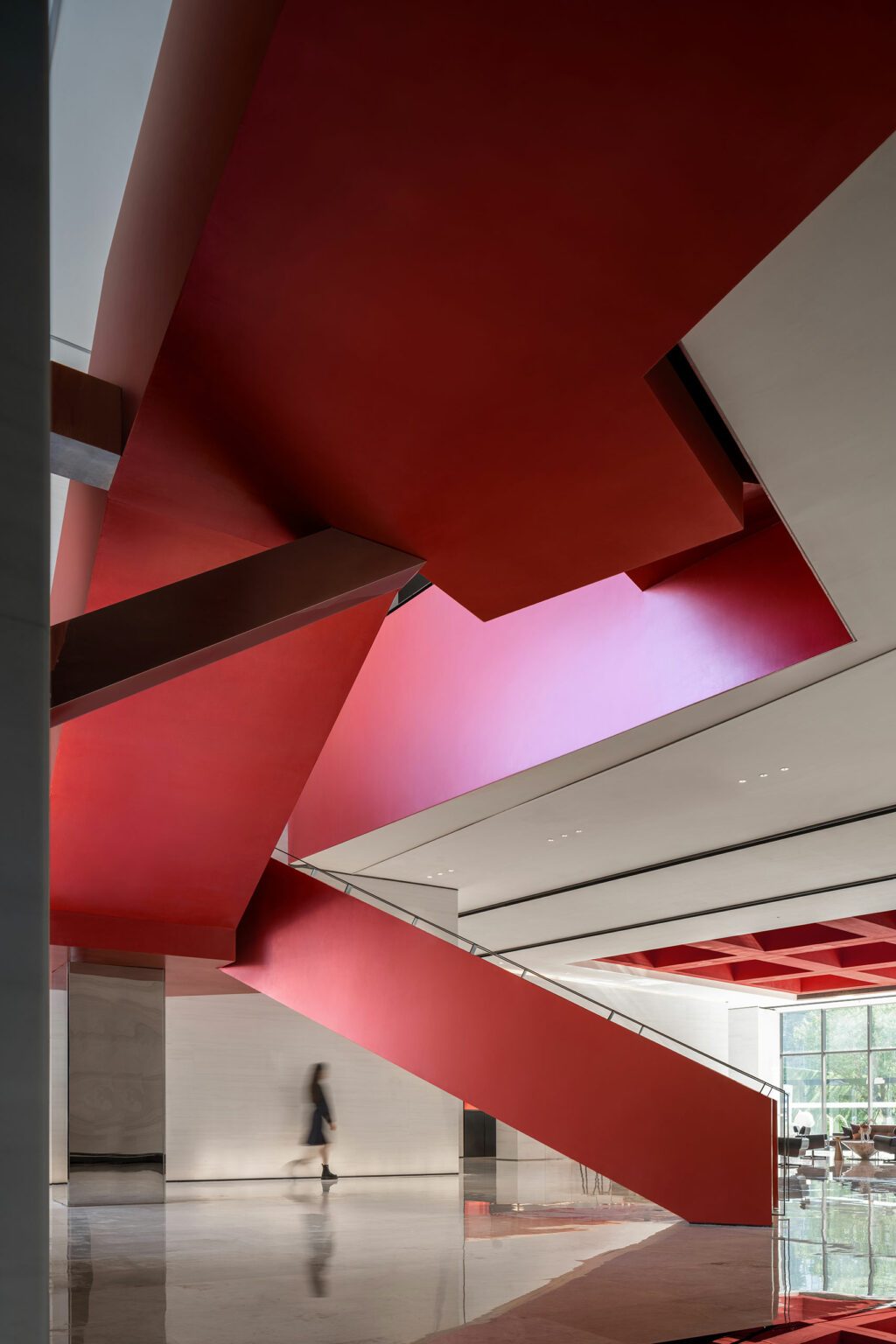 A striking red staircase with modern geometric design dominates a spacious, minimalist lobby. A blurred figure walks below, adding a dynamic element.