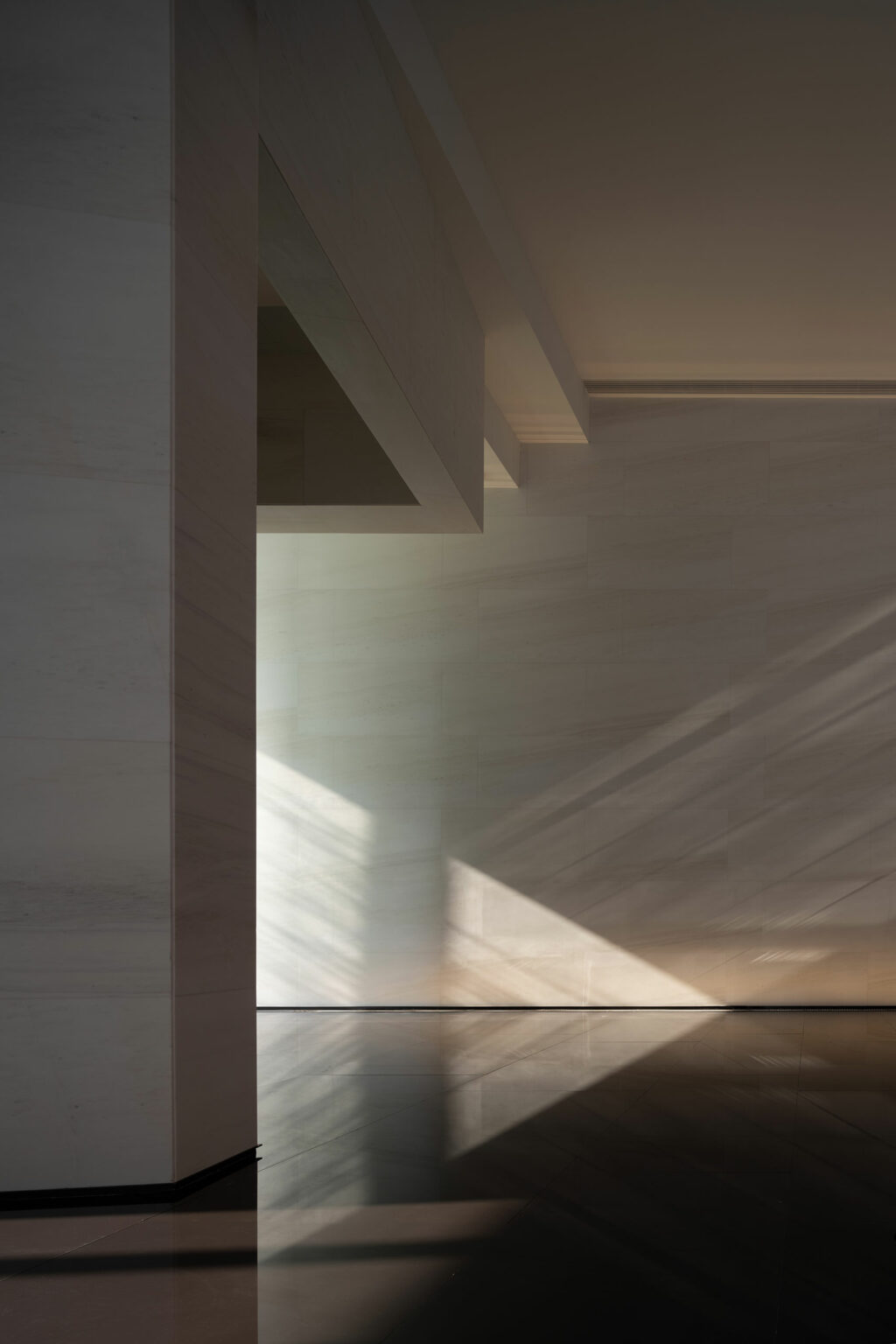 Minimalist interior with sunlight casting geometric shadows on smooth, light-colored walls and reflective dark flooring. A calm, serene atmosphere.