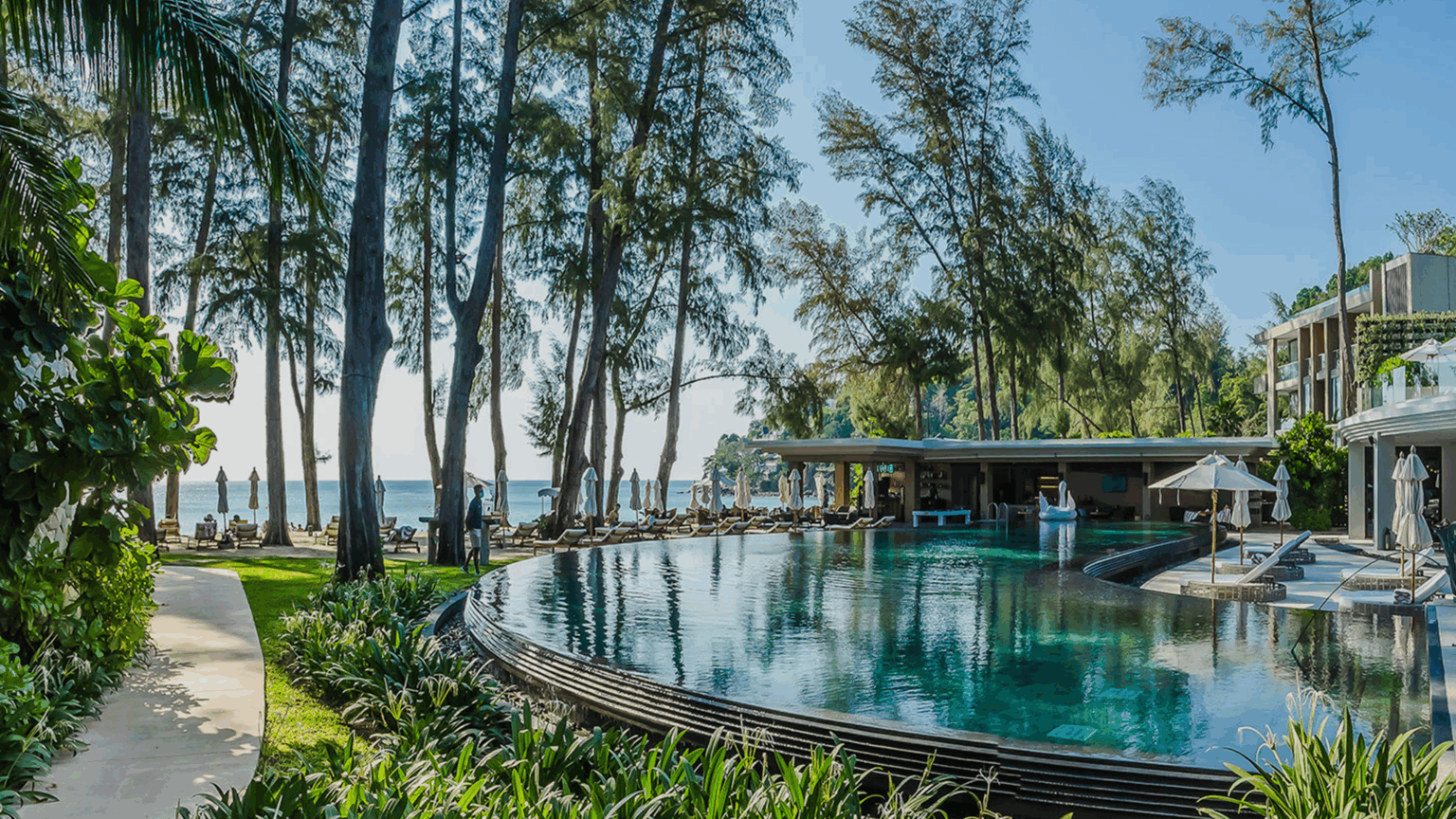 Luxurious resort view with an infinity pool surrounded by lush greenery, sun loungers, and large trees. The ocean is visible beyond, under a clear blue sky.
