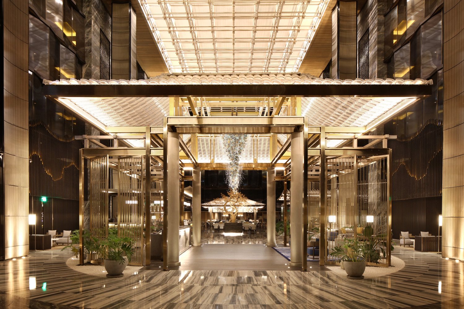 Luxurious hotel lobby featuring a grand golden portal, sculptural chandelier, statement tree installation, and integrated lighting under a vaulted glass ceiling.