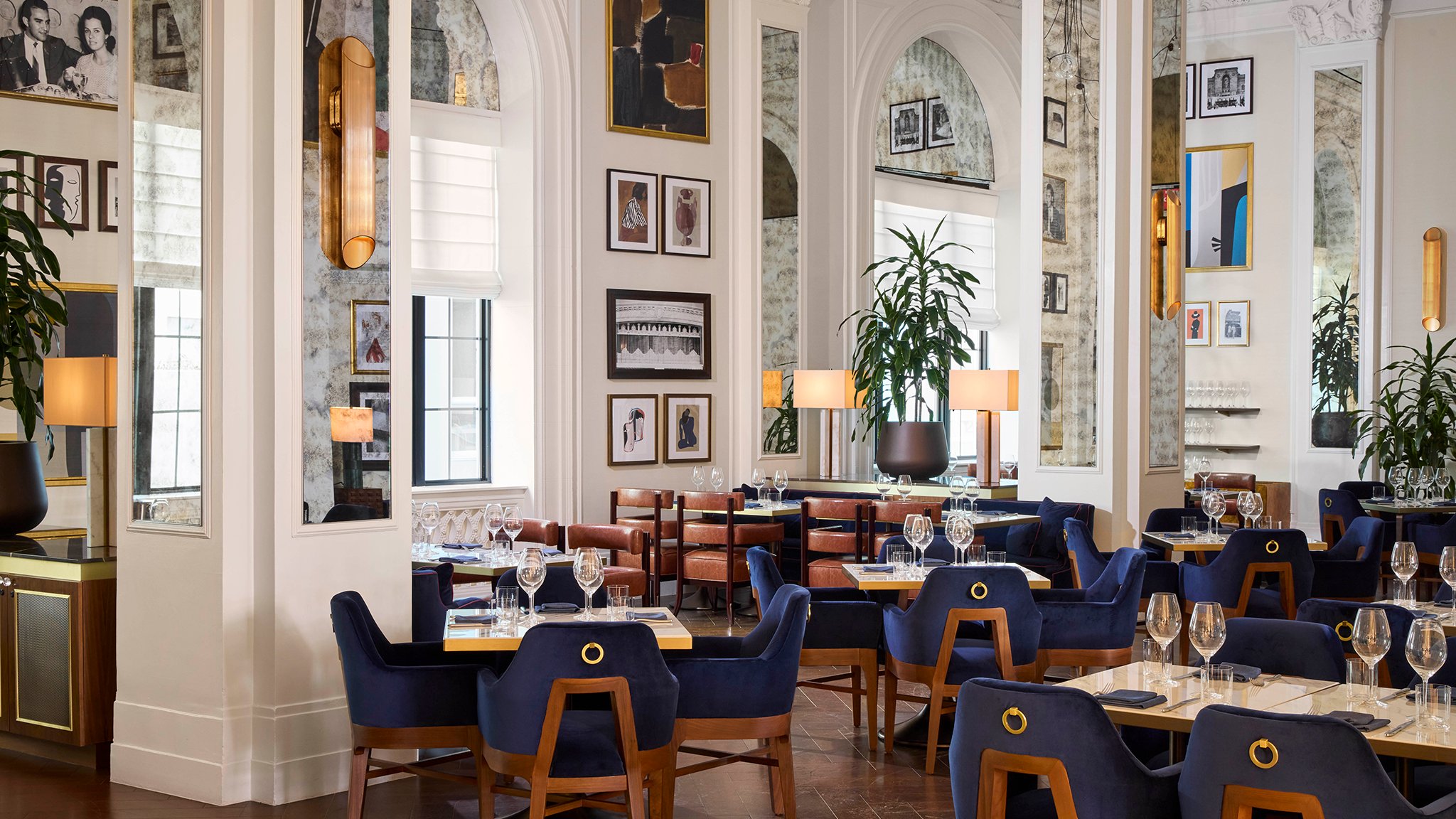 Elegant restaurant interior with blue velvet chairs, wooden tables, and framed artworks on mirrored walls. Warm lighting and lush plants create a cozy ambience.