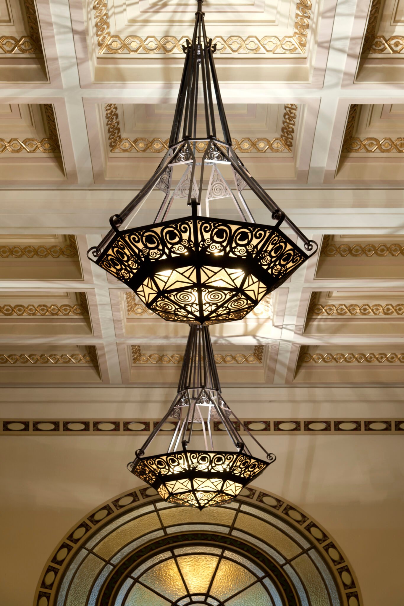 Ornate chandeliers with intricate ironwork and stained glass illuminate a ceiling adorned with gold accents and geometric patterns, exuding elegance.