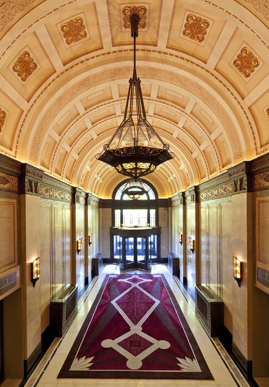 Ornate lobby with arched ceiling, elegant chandelier, and geometric maroon carpet. Features warm lighting and a large revolving glass door.