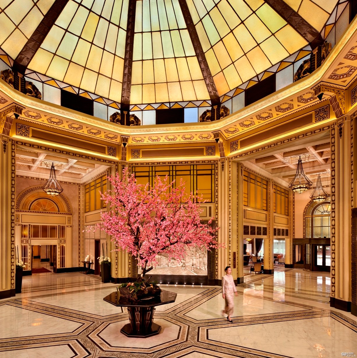 Luxurious hotel lobby adorned with a large, blooming pink cherry blossom tree. Elegant woman in traditional attire walks beneath a grand stained-glass dome.