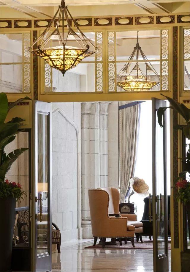 Elegant hotel lobby with ornate chandeliers, vintage armchairs, an old phonograph, and large windows draped with curtains. The ambiance is classic and inviting.