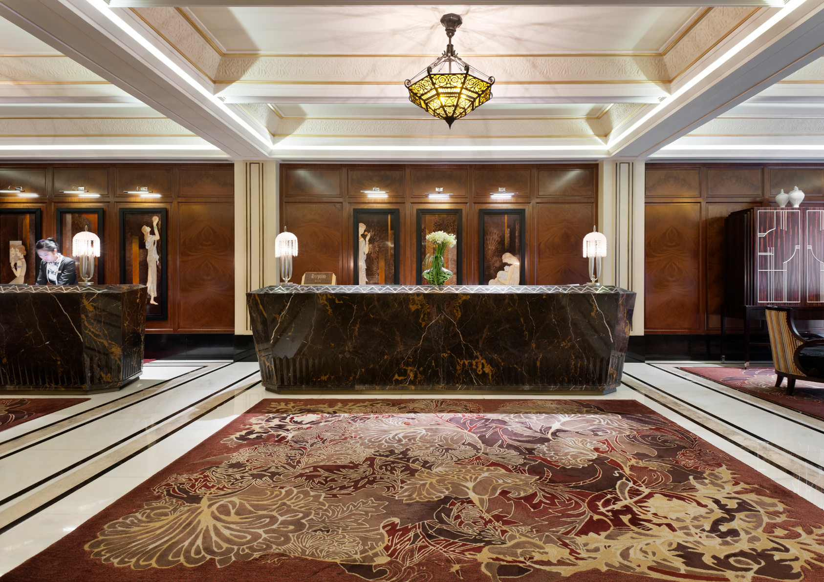 Elegant hotel reception area with a dark marble reception desk, ornamental lamps, floral arrangements, decorative wooden panels, and a plush patterned carpet.