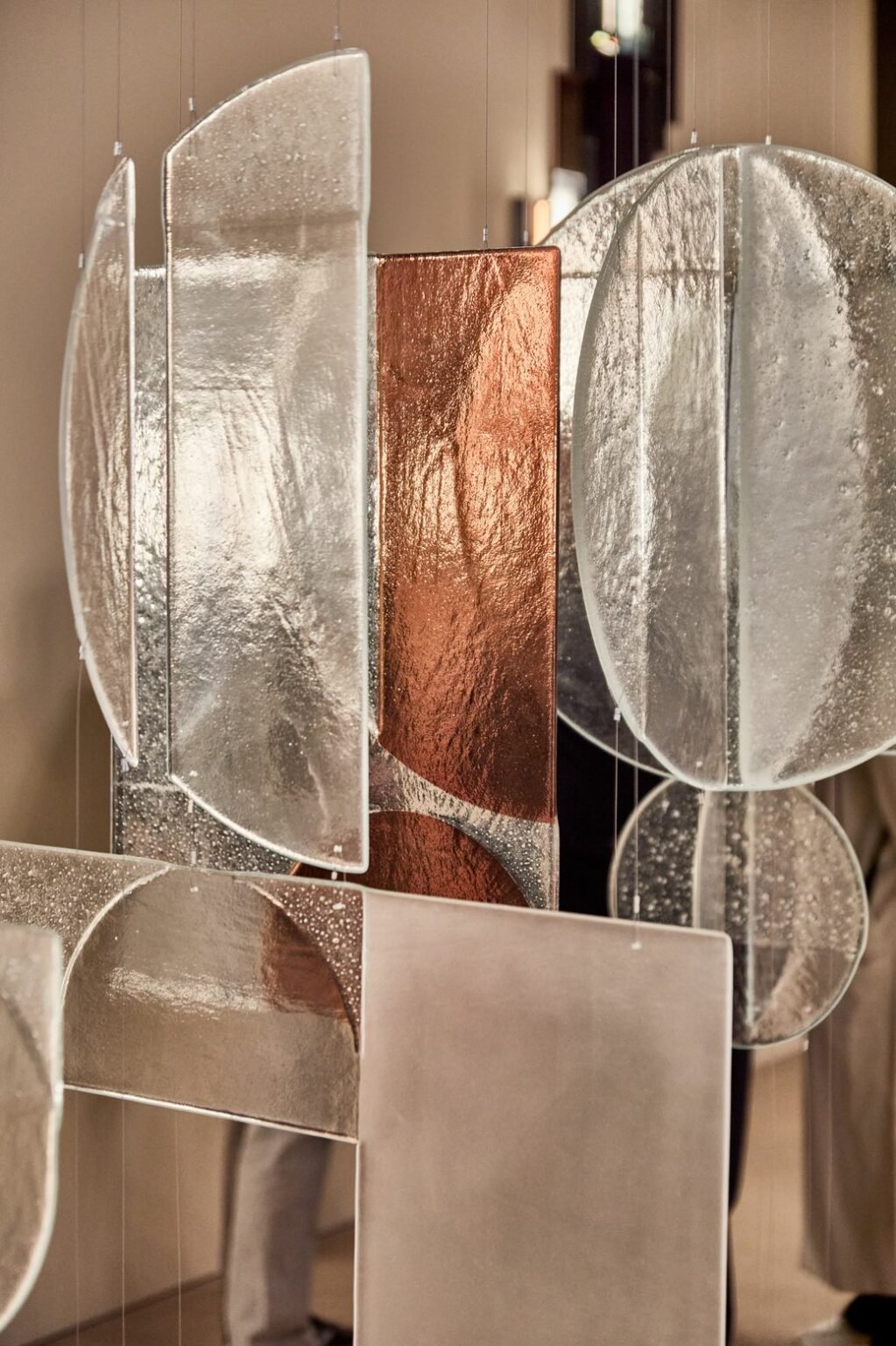 Hanging modern sculptures featuring rectangular and circular textured glass in silver and copper tones. Reflective surfaces and elegant lines create a sophisticated atmosphere.