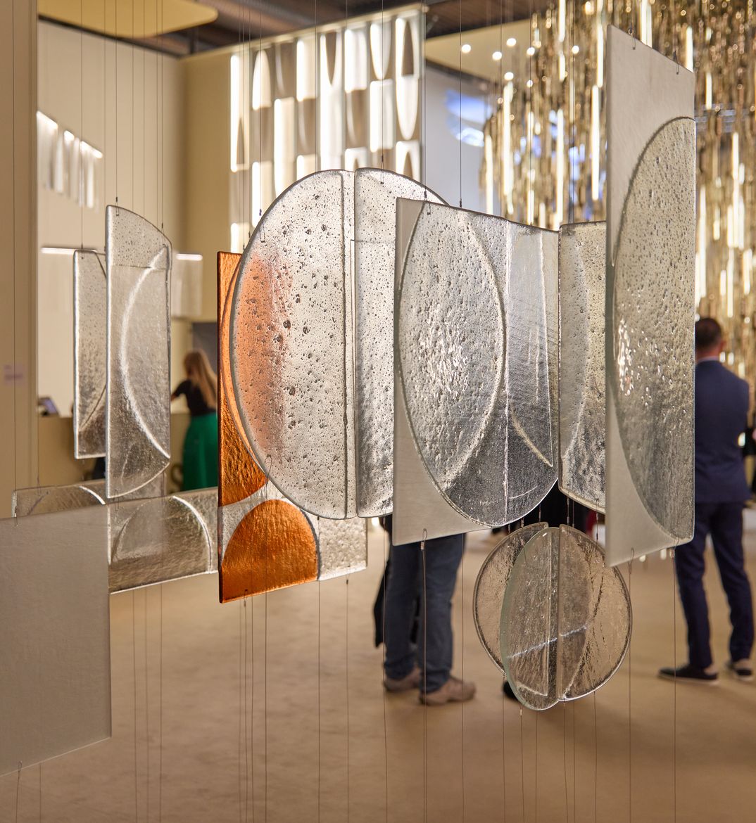 Hanging glass panels with textured circular patterns, some in orange and others clear, create a modern art installation. People observe in the background.Hanging glass panels with textured circular patterns, some in orange and others clear, create a modern art installation. People observe in the background.