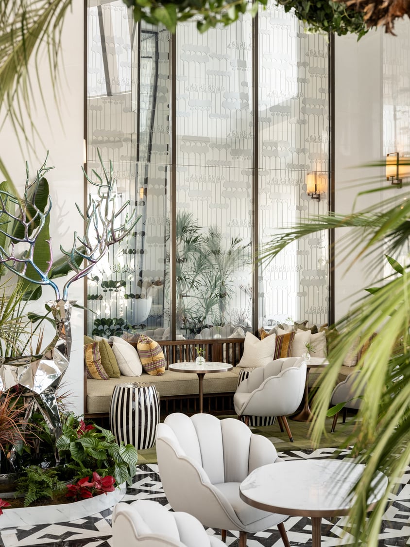 Luxurious lounge with white armchairs and round tables, surrounded by greenery and patterned walls. Mirrors and plants add an elegant, serene feel.