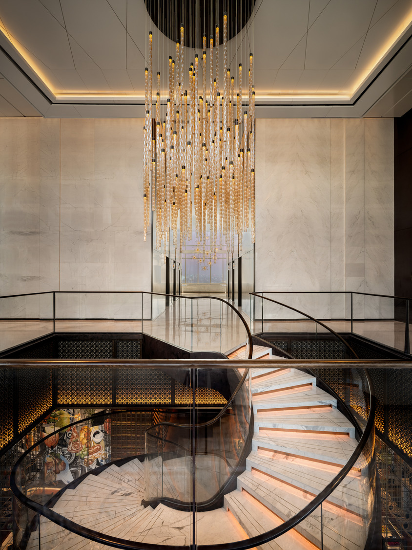 Elegant interior with a spiral marble staircase and glass railing. Above, a large, intricate chandelier with hanging lights exudes a luxurious ambiance.