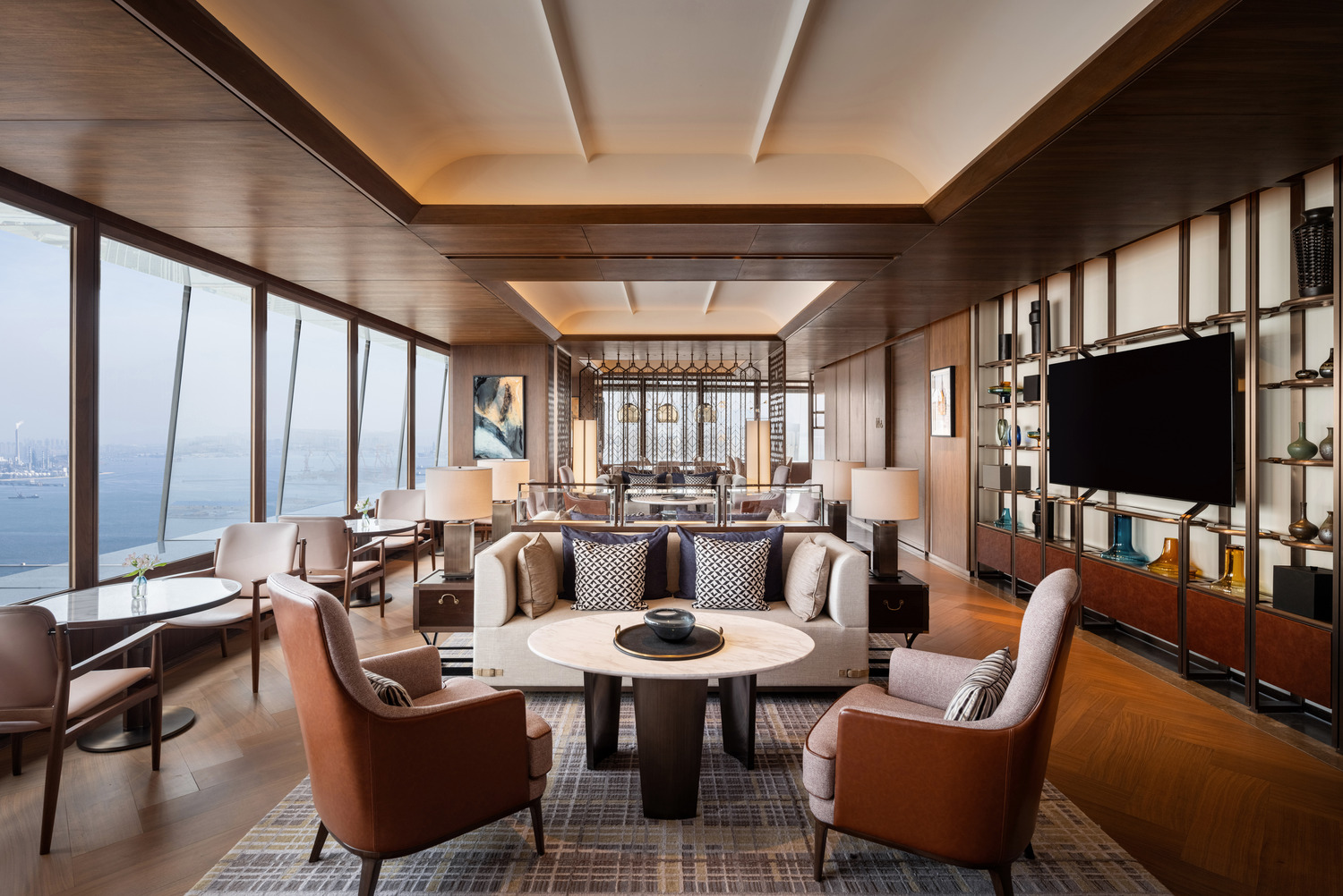 Luxurious lounge with ocean view, featuring plush beige sofas, brown leather chairs, and a round marble table. Elegant decor with wood accents.