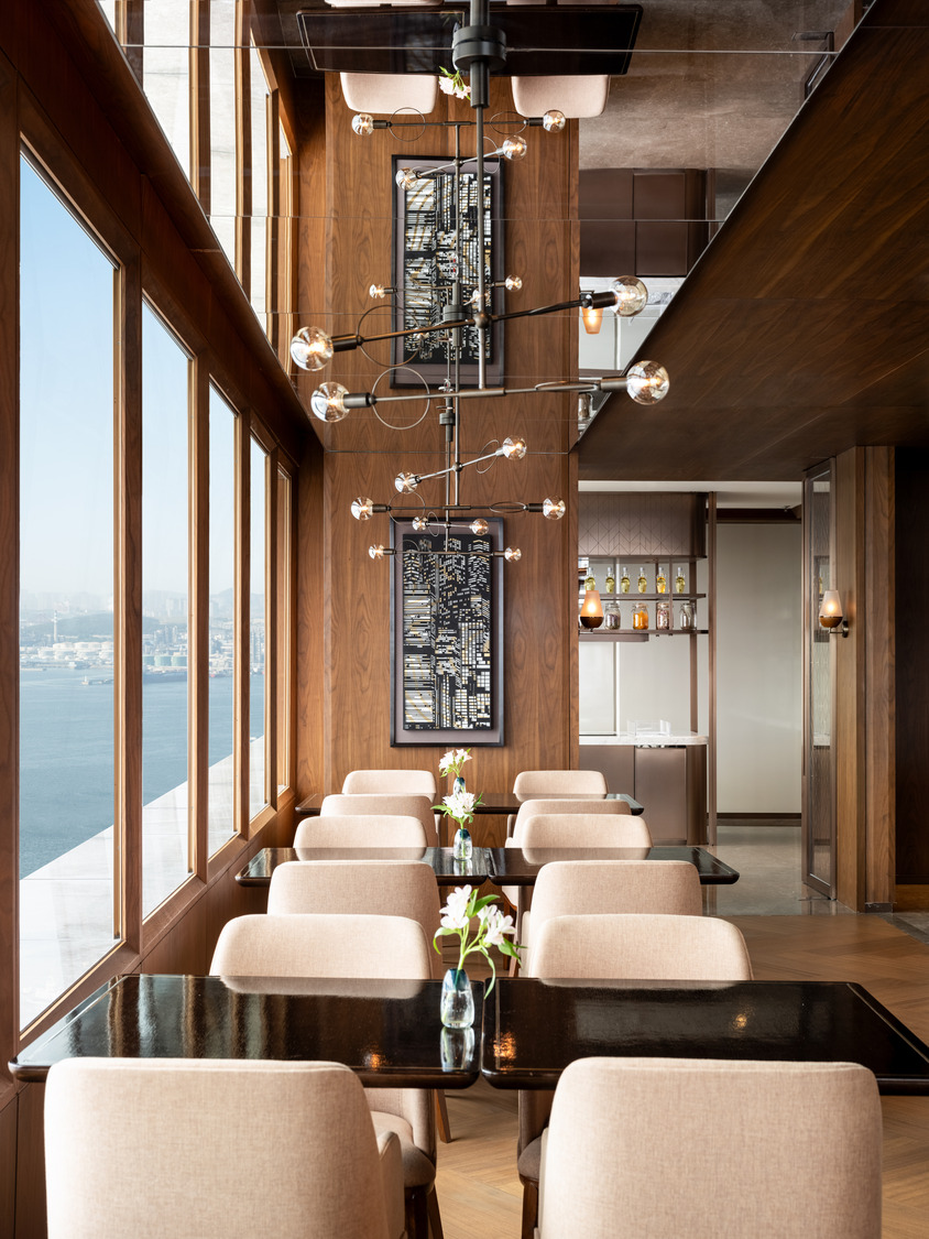 Modern restaurant interior with beige chairs and black tables set with flowers. Large windows offer city and ocean views. Warm wood paneling and a chic chandelier create an elegant ambiance.