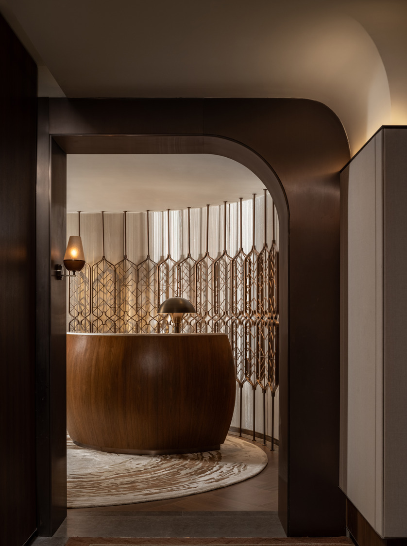 A modern interior with a curved wooden reception desk, a stylish lamp, and a geometric screen divider. Warm tones create an inviting, elegant atmosphere.