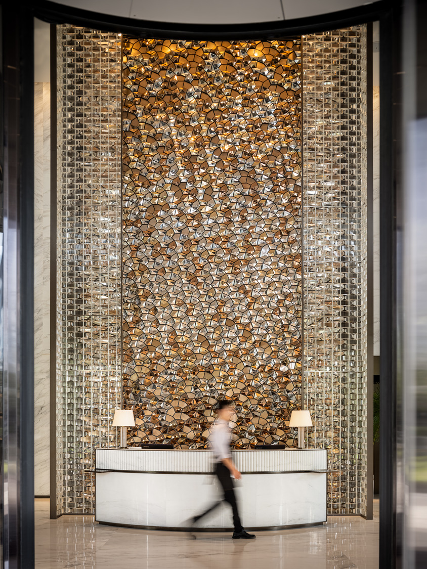 A man walks by a luxurious reception desk with two lamps, set against a stunning, reflective mosaic wall with gold and silver patterns, exuding elegance.