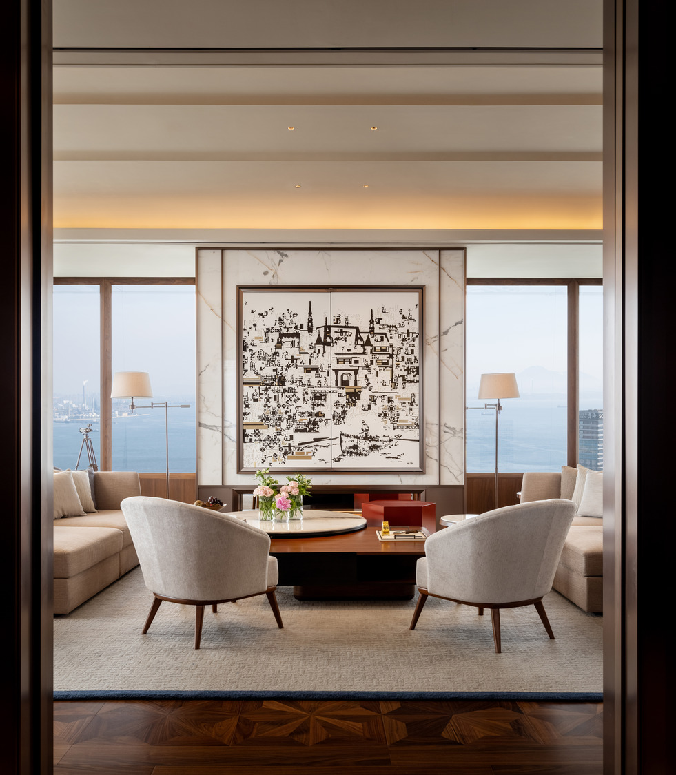 Elegant living room with beige armchairs, a wooden coffee table, and a large black-and-white artwork. Floor-to-ceiling windows offer a sea view.