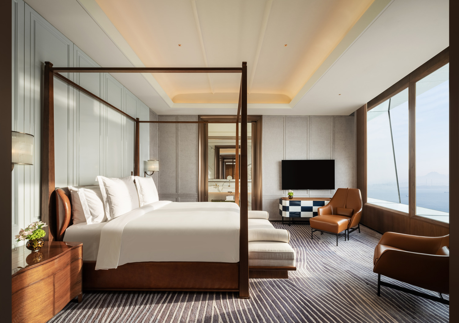Luxurious hotel room with a four-poster bed, brown leather armchairs, and wide ocean-view windows. Neutral tones create a serene atmosphere.