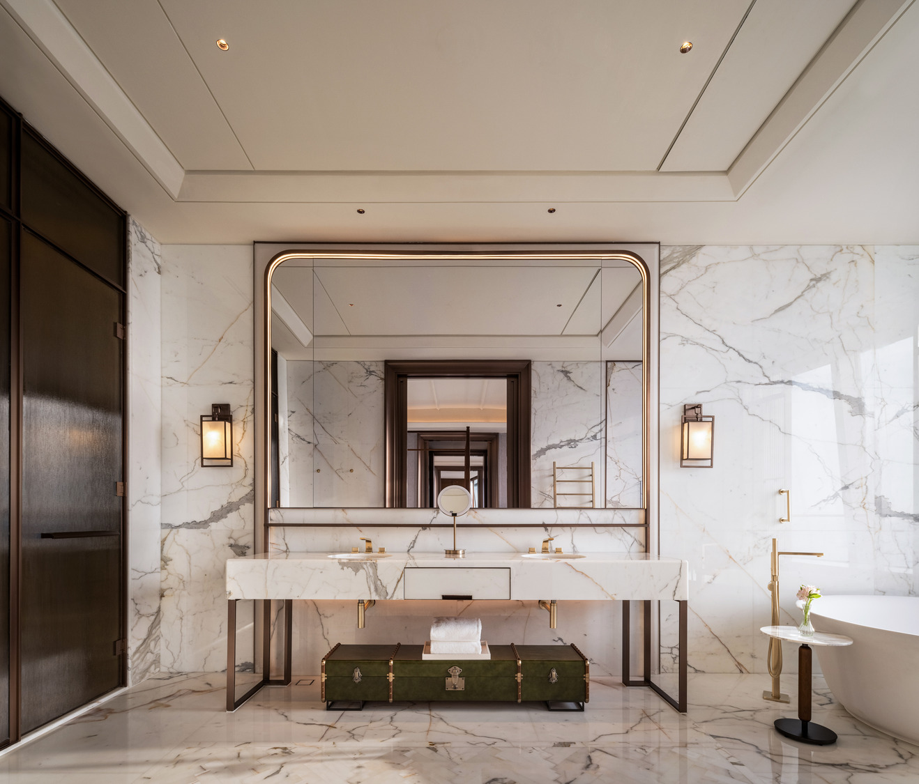 Luxurious marble hotel bathroom with a large mirror, elegant lighting, and gold accents. A green trunk sits under the sink, while a vase with flowers decorates a small table.