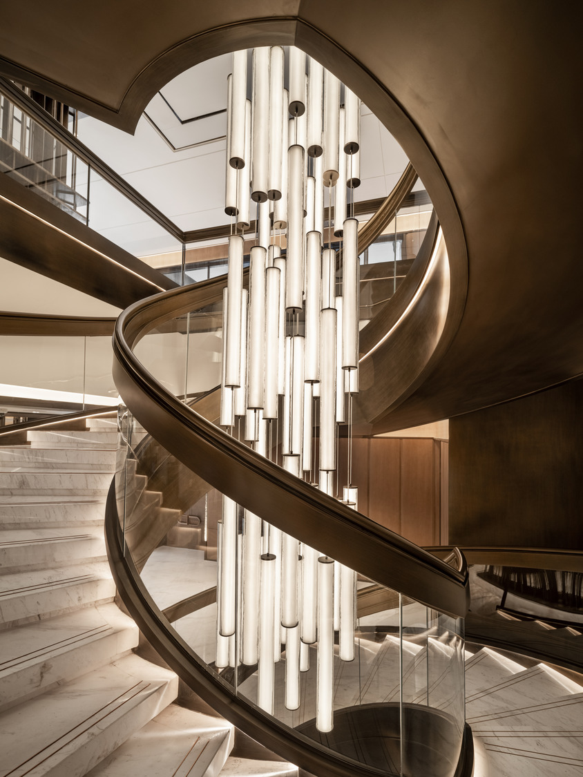 Elegant spiral staircase with marble steps and glass railing, illuminated by vertical tubular lights. The setting is modern and sophisticated.