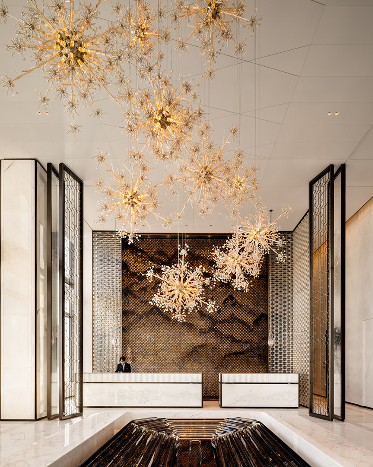 Elegant hotel lobby with large, gold starburst chandeliers hanging from high ceiling. Two reception desks below a textured mosaic wall, creating a luxurious ambiance.
