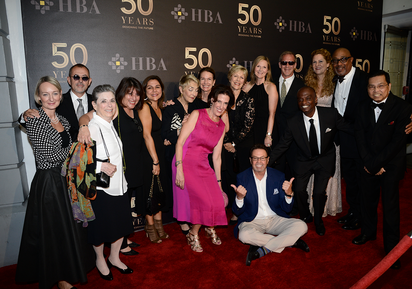 A diverse group of designers in formal attire pose joyfully on a red carpet, celebrating
