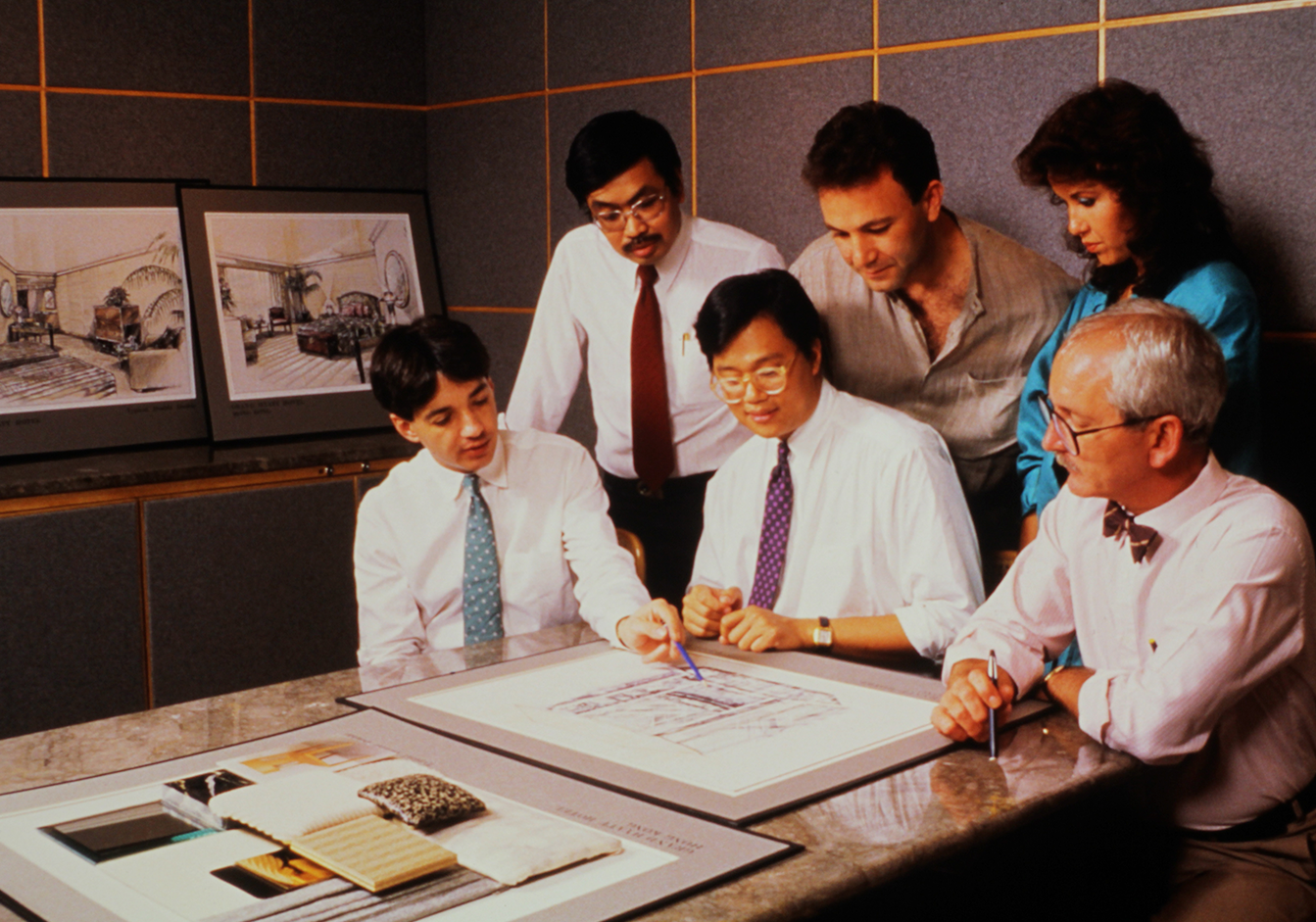 A group of six designers from HBA Hong Kong studio gathers around a table, intently reviewing architectural sketches. The mood is focused and collaborative, emphasizing teamwork in design.