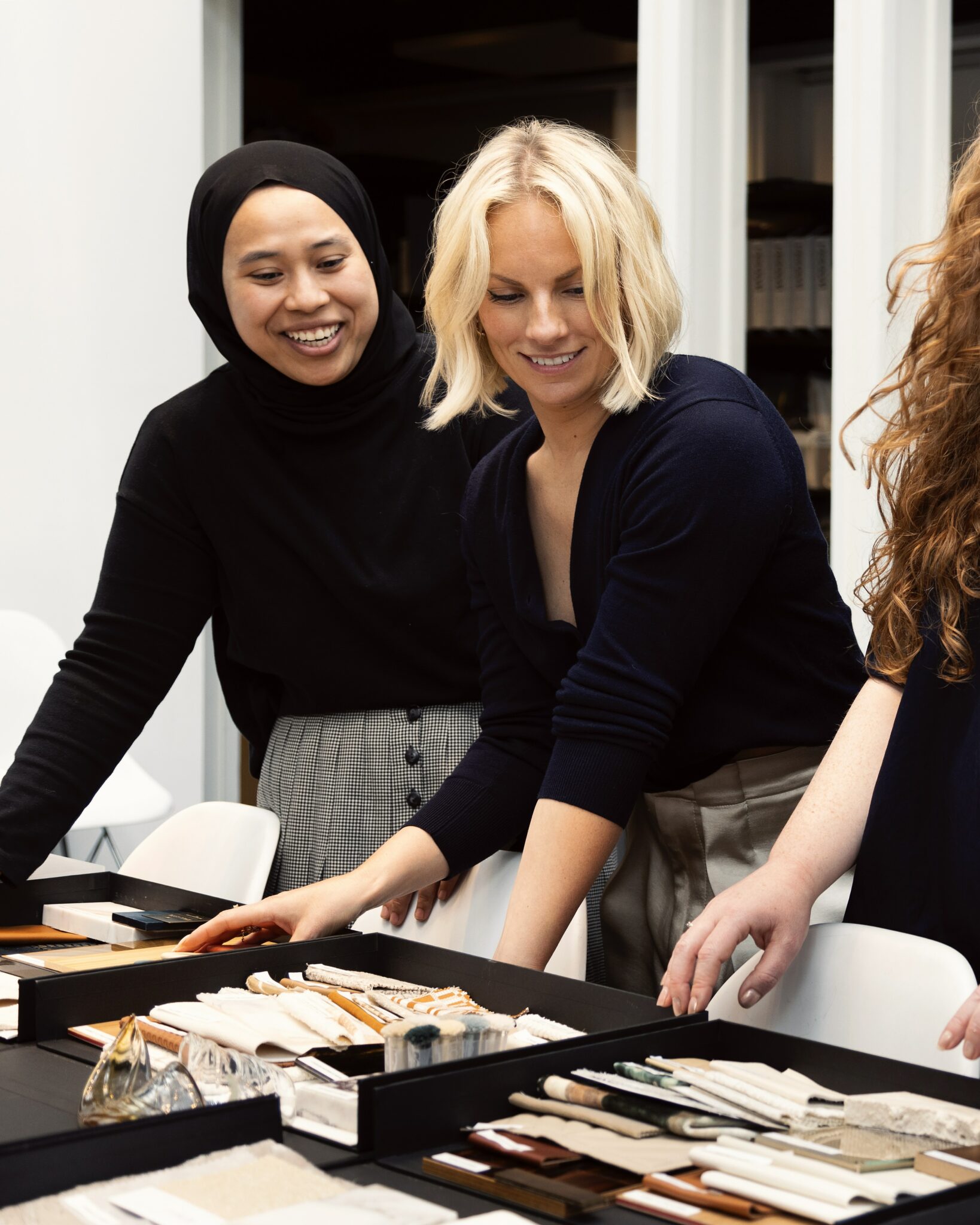Two young female designers smiling and leaning over a table with fabric samples and moodboards in HBA London studio. They appear engaged and collaborative, conveying a positive, creative atmosphere.