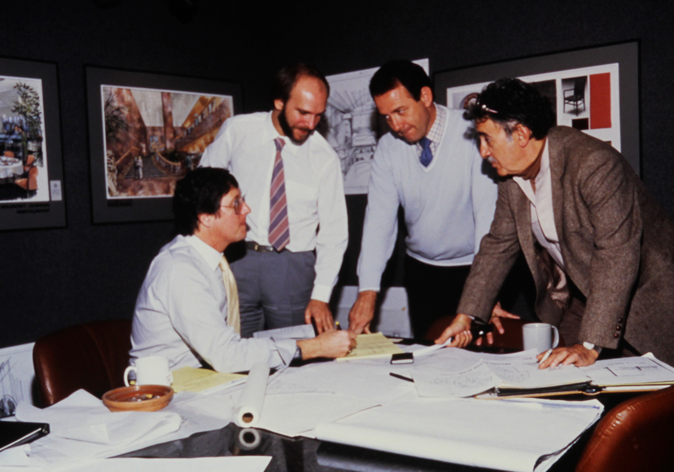 Four designers from HBA Los Angeles studio in business attire engaged in a discussion around a table cluttered with blueprints and papers, conveying a serious and collaborative atmosphere.