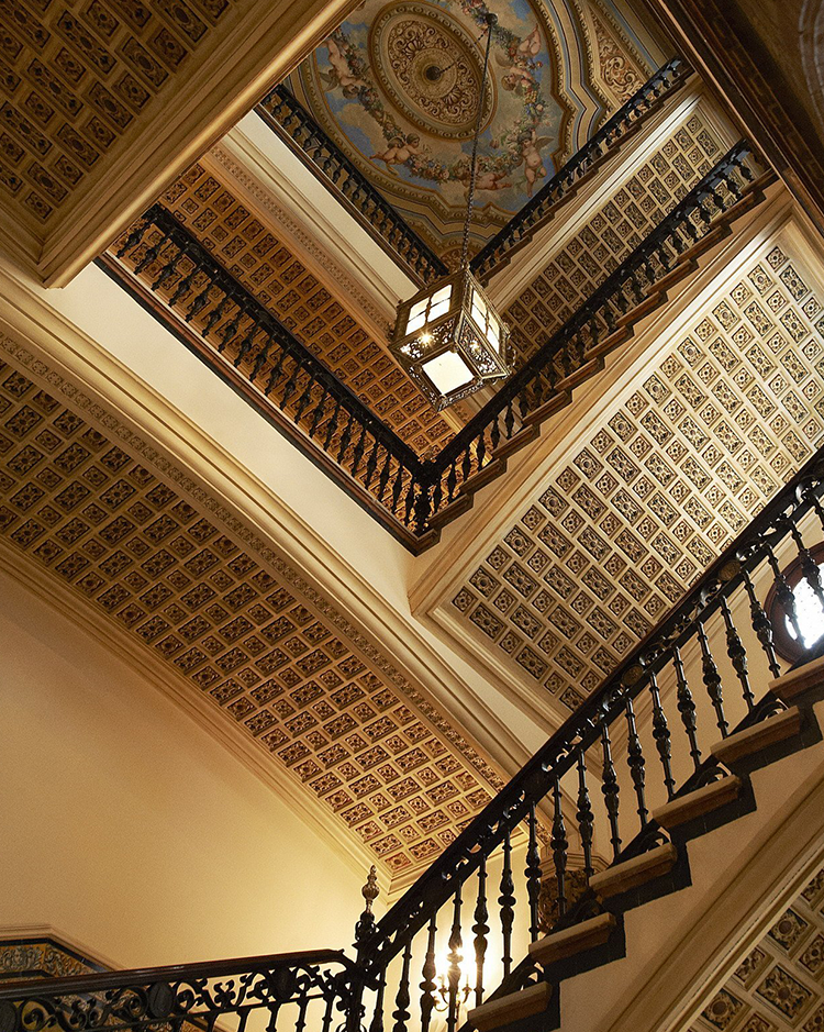 Ornate stairwell with intricate patterned ceilings and a decorative hanging lantern. Rich textures and elegant railings create a grand, historic atmosphere.