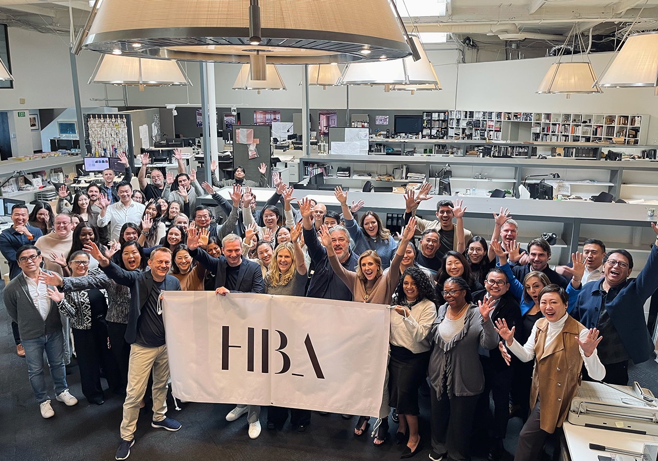 A large, diverse group of smiling designers from in the HBA London studio raise their arms in celebration, holding an