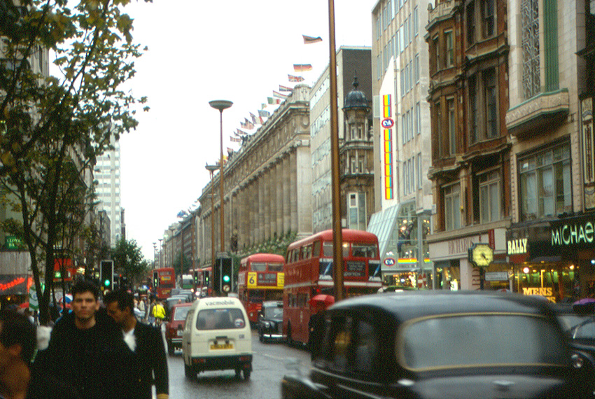London's busy street with red double-decker buses and cars on a rainy day. Buildings line the road, adorned with flags. People walk on sidewalks, conveying urban bustle.