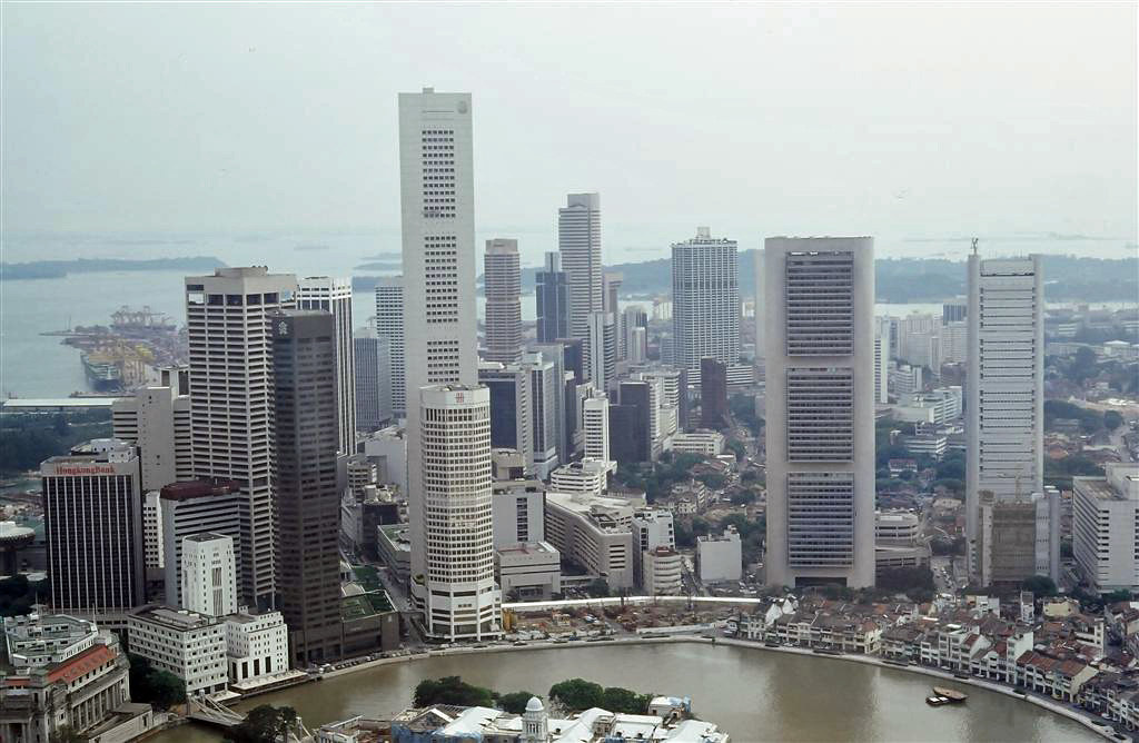 Aerial view of Singapore's bustling cityscape with tall, modern skyscrapers. The buildings surround a winding river, and a shipyard is visible in the background. The scene conveys urban vibrancy.