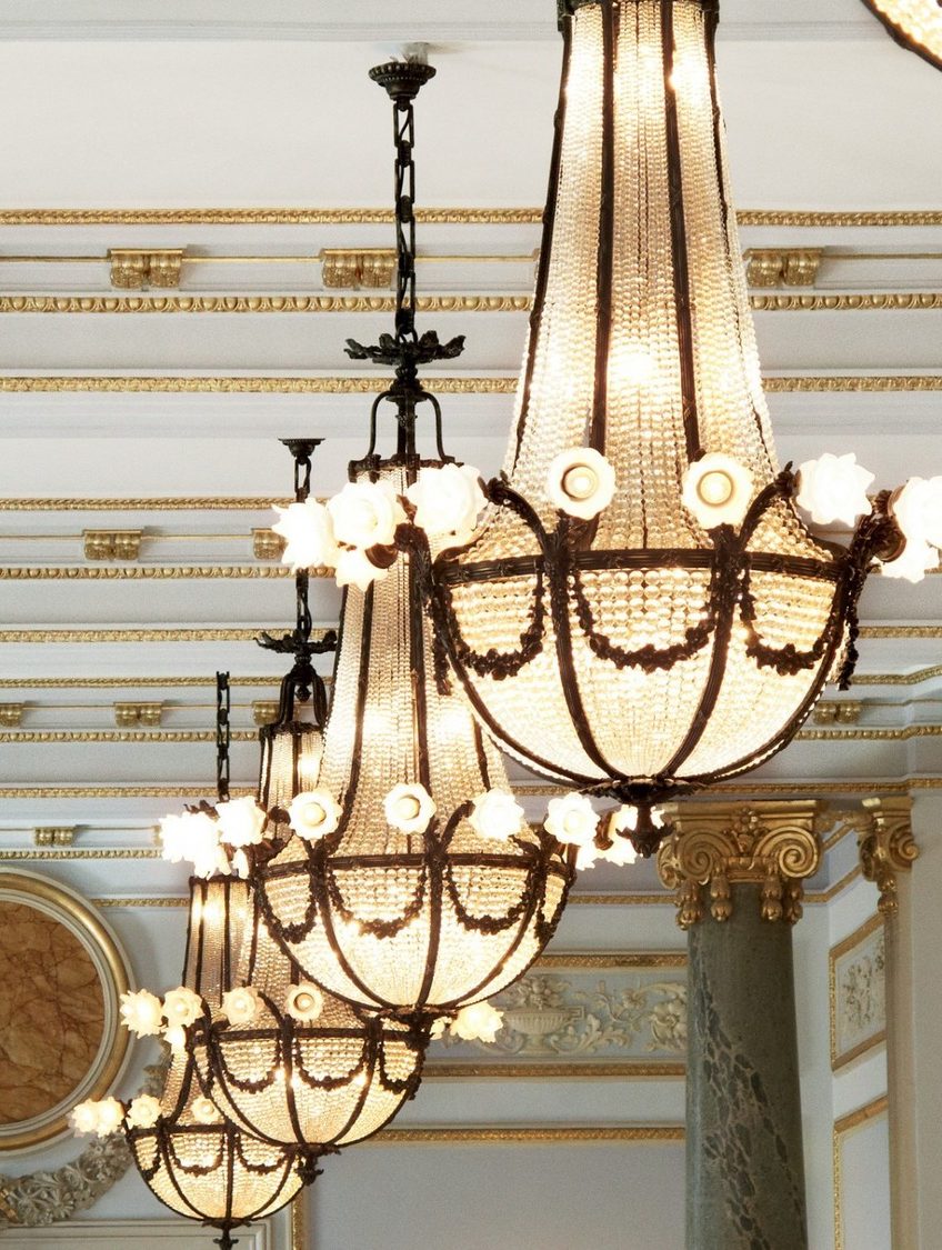 Elegant chandeliers with crystal details hang from an ornate ceiling with gold accents and intricate molding, exuding a luxurious ambiance.