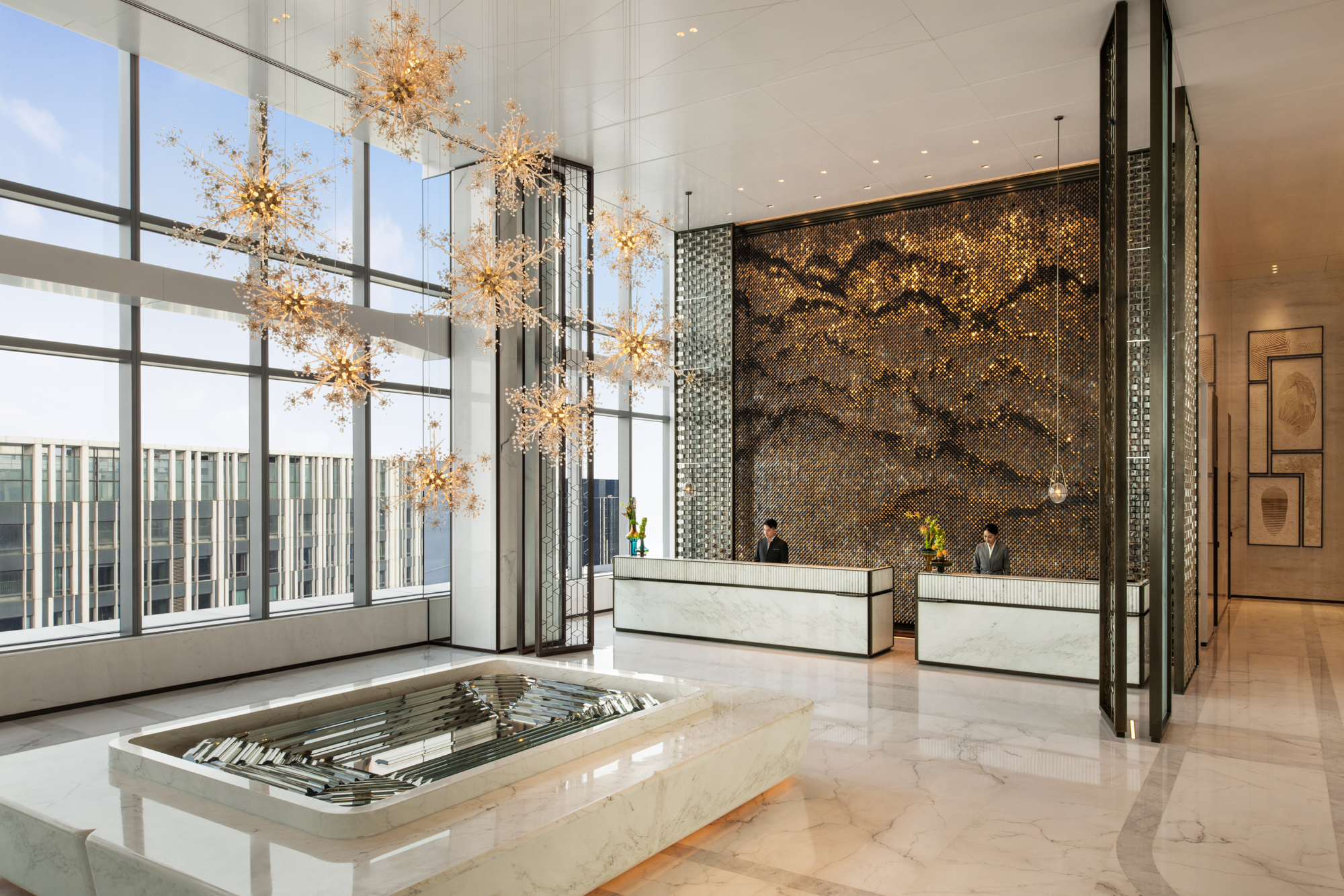 Elegant hotel lobby with large, gold starburst chandeliers hanging from high ceiling. Two reception desks below a textured mosaic wall, creating a luxurious ambiance.