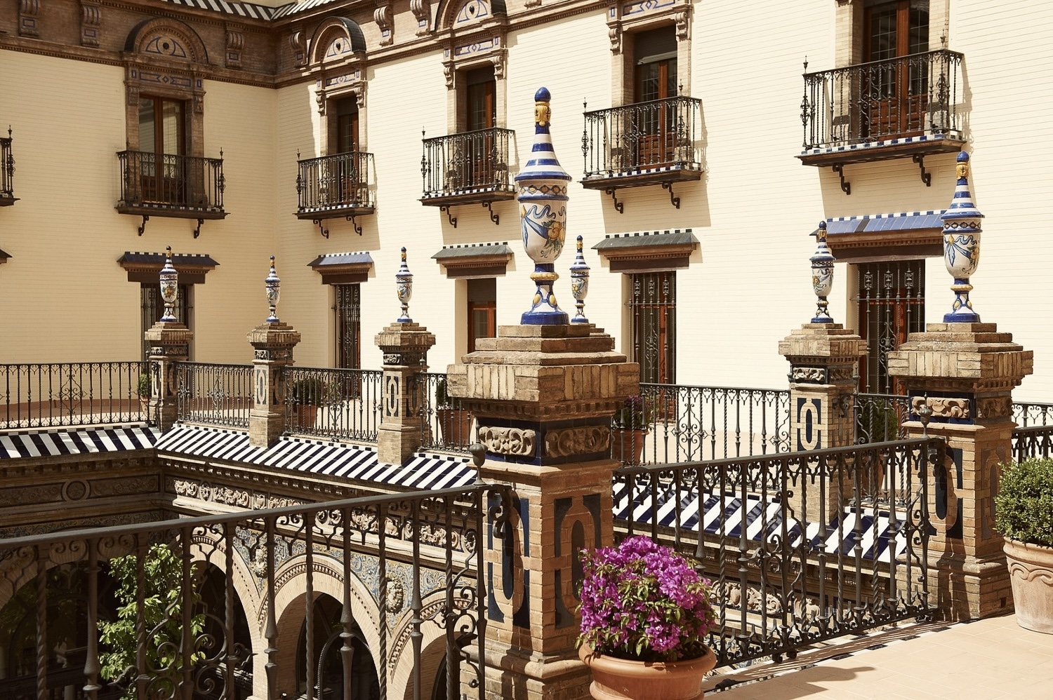Exterior of Hotel Alfonso XIII, a Luxury Collection Hotel, Seville, Spain. Elegant courtyard with ornate iron railings and decorative ceramic urns atop brick pillars. Balconies feature striped canopies and potted purple flowers, creating a serene, historic ambiance.