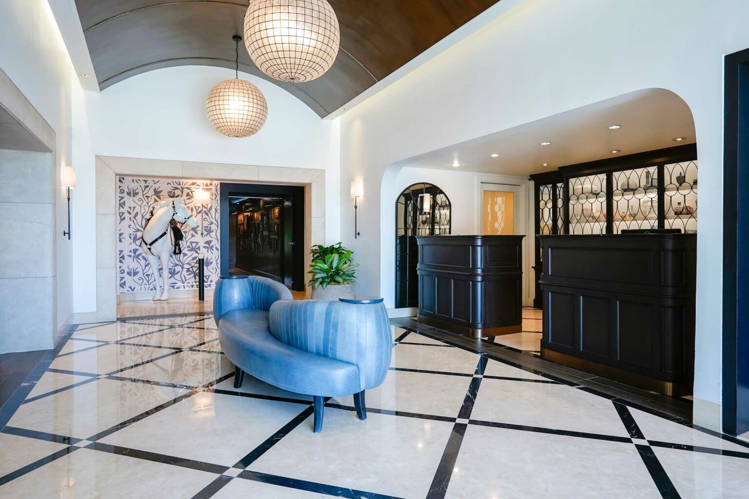 Elegant hotel lobby with a curved blue sofa, two spherical chandeliers, black reception desks, and horse sculpture on a patterned wall, conveying sophistication.