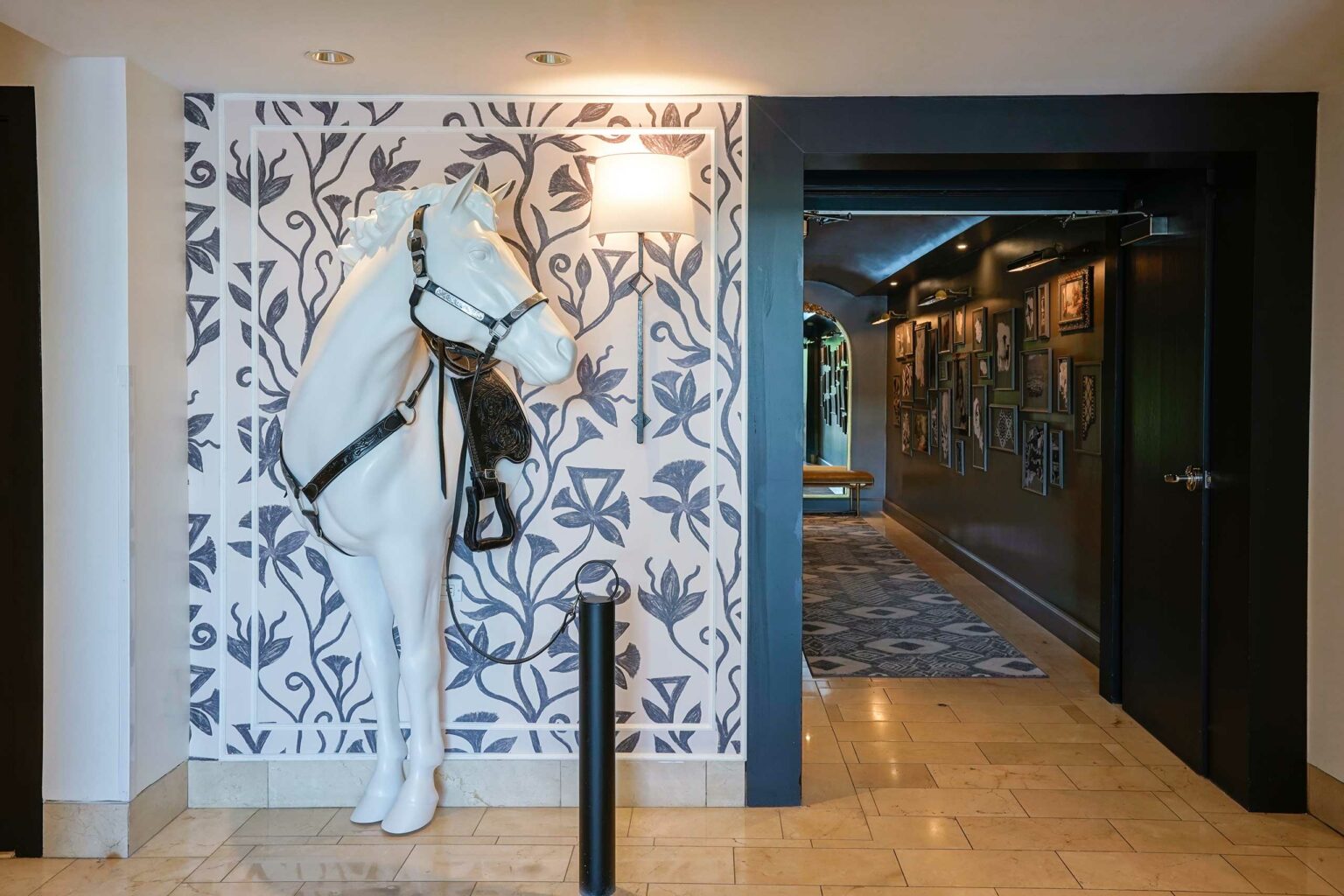 A white horse statue with a black harness stands against a floral-patterned wall in a hotel hallway. The decor is modern with a gallery of photos visible.