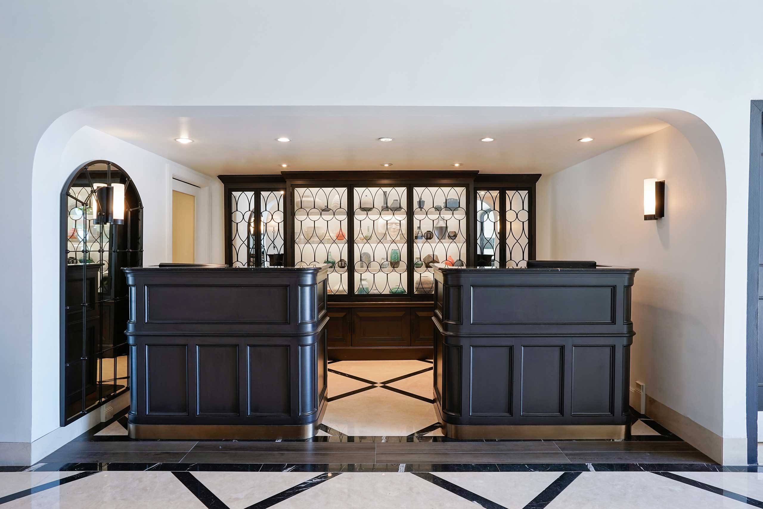 Spacious hotel lobby with two dark wooden reception desks under soft lighting. Glass display shelves behind showcase colorful glassware, creating an elegant ambiance.