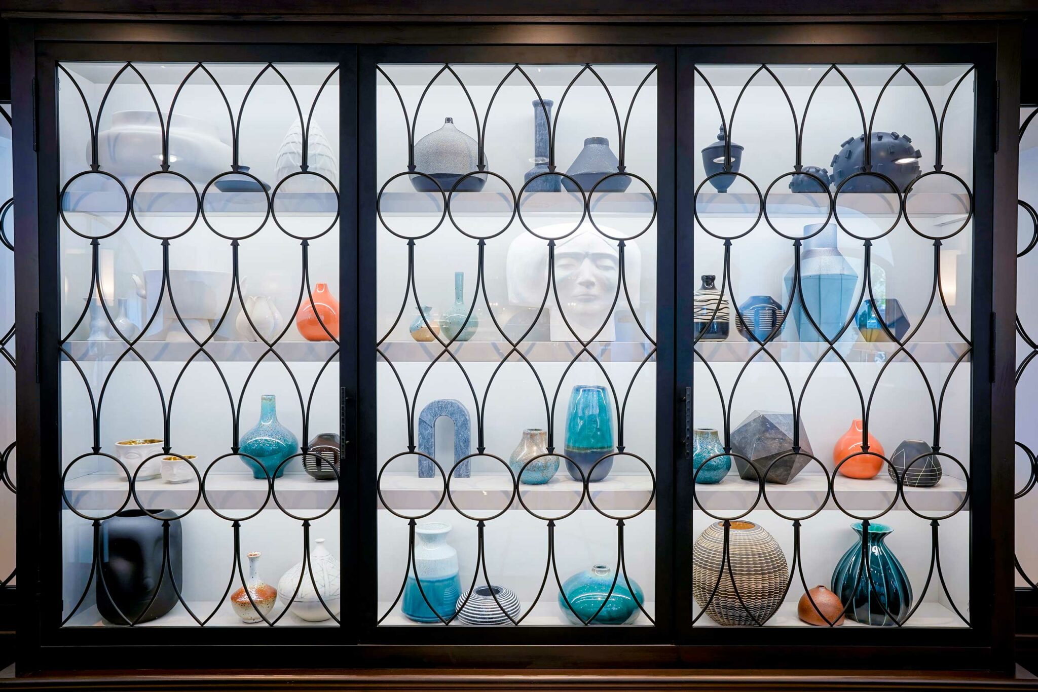 A glass cabinet displays colorful, diverse pottery and vases, with shades of blue, orange, and black, behind decorative black ironwork, creating an artistic and elegant atmosphere.