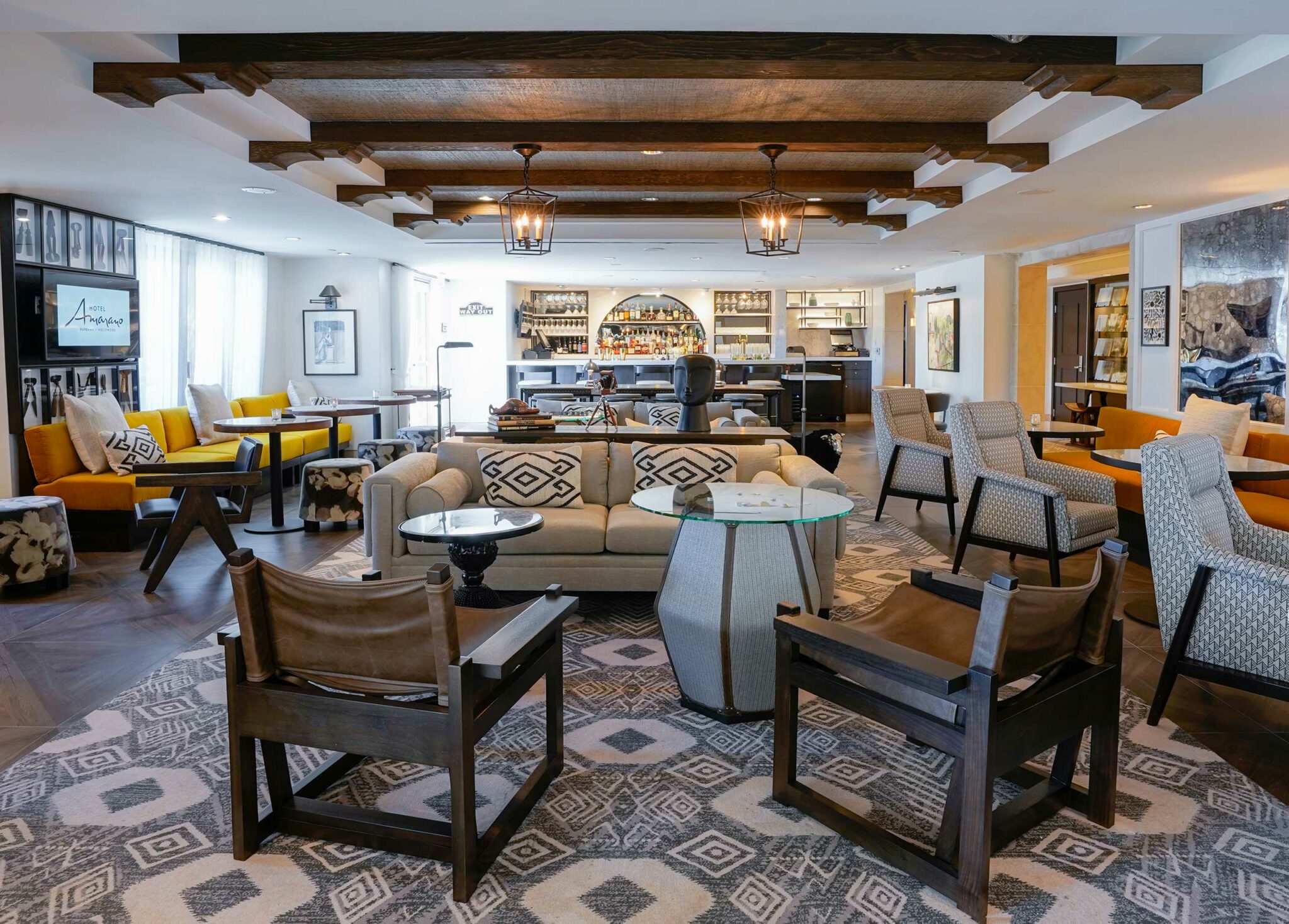 Cozy hotel lounge with patterned couches, wooden chairs, and vibrant yellow accents. Warm lighting from ceiling lanterns and a stocked bar in the background.