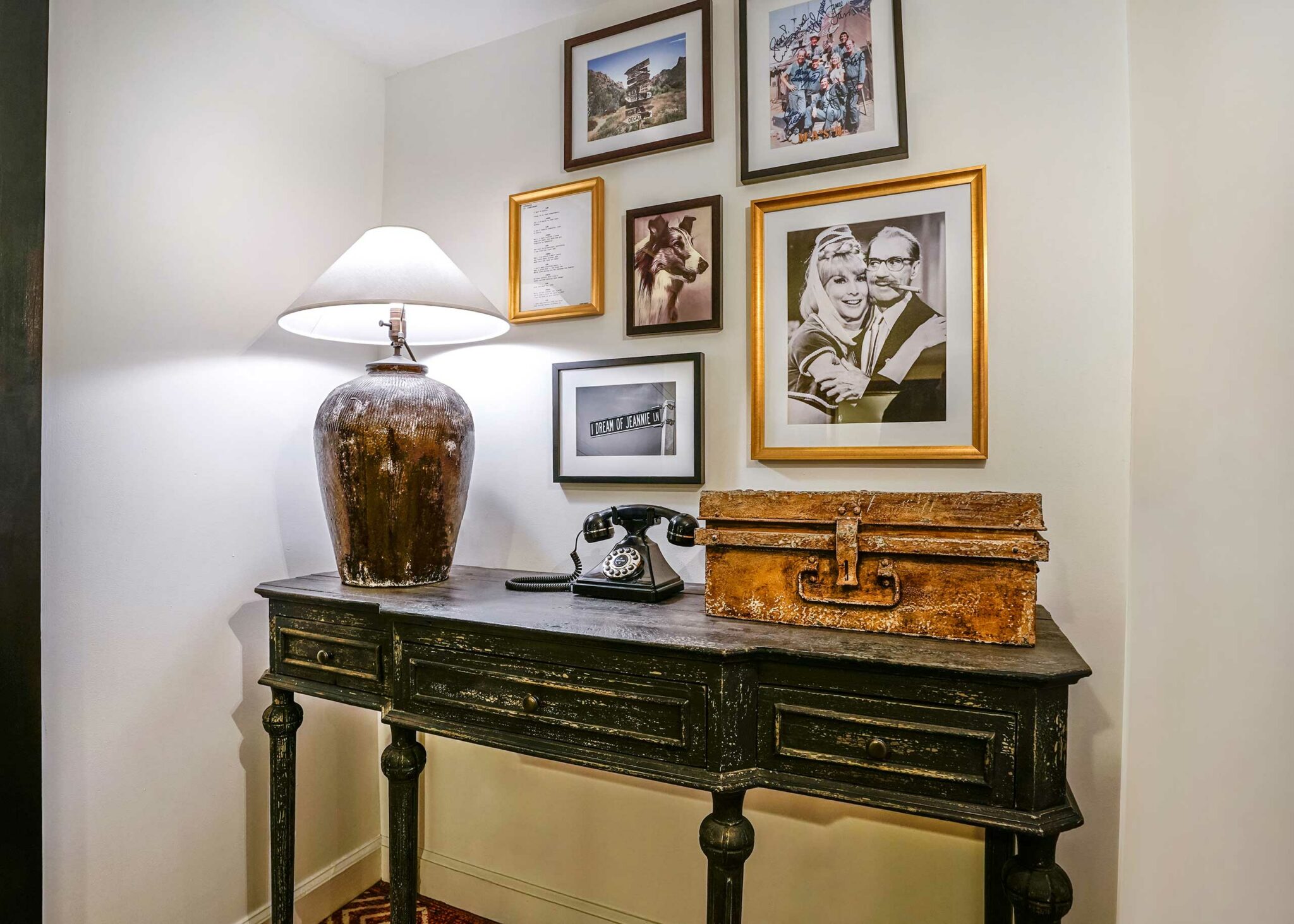A vintage desk holds a large lamp, retro telephone, and a rustic box. Above, a gallery wall features framed photos and art, creating a nostalgic atmosphere.