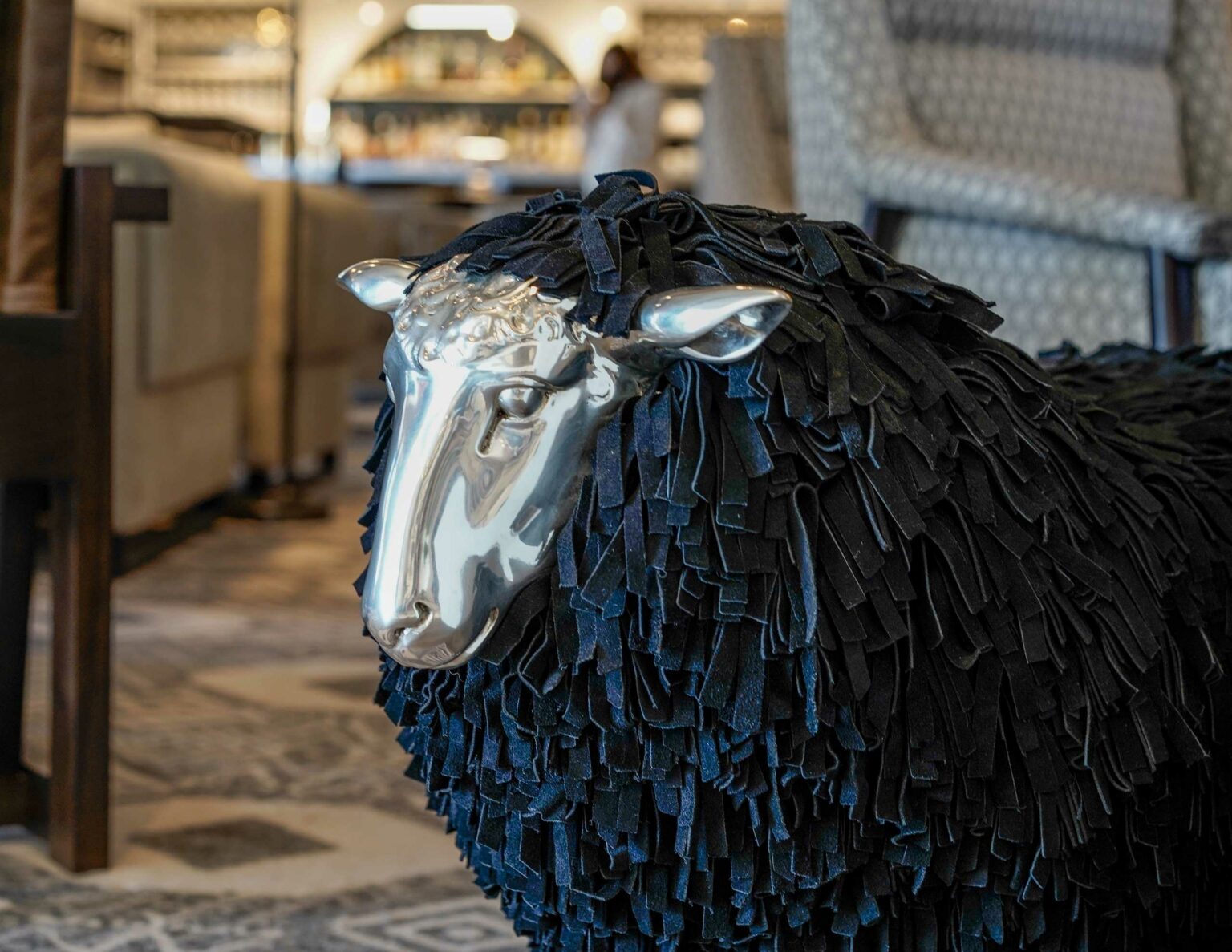 A sculpture of a black sheep with a shiny metallic face and ears is displayed indoors. The body is textured with dark fringe, creating an elegant contrast.