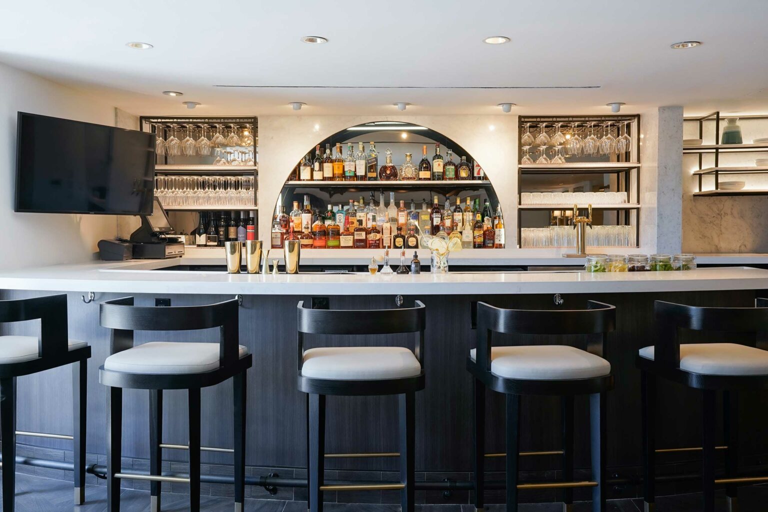 Sleek modern hotel bar with dark wood and white accents, lined with empty stools. Shelves display various liquor bottles and glassware, creating an inviting atmosphere.
