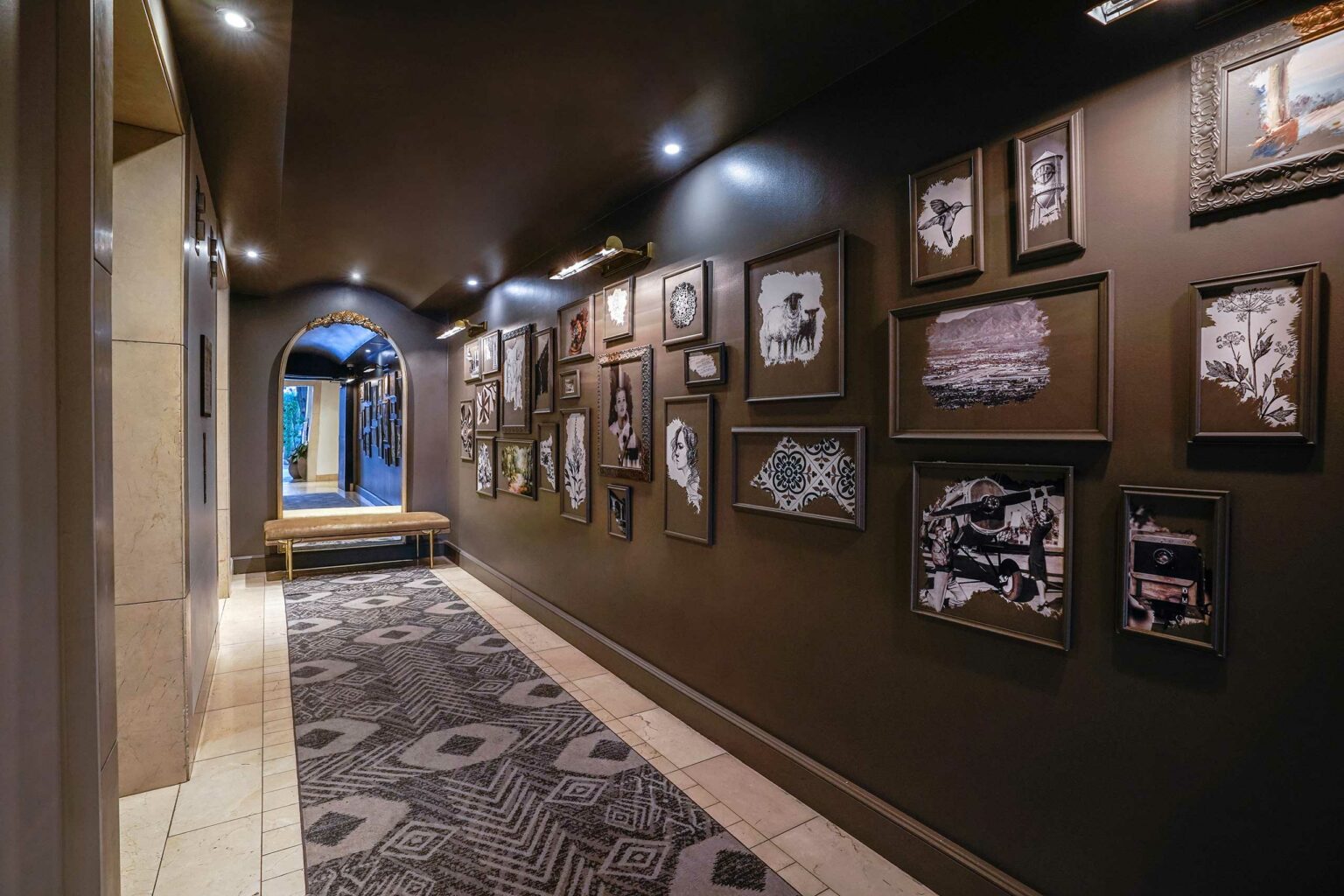 Dimly lit, elegant hotel hallway with dark walls, featuring an eclectic gallery of framed art. An archway and a patterned rug add sophistication.