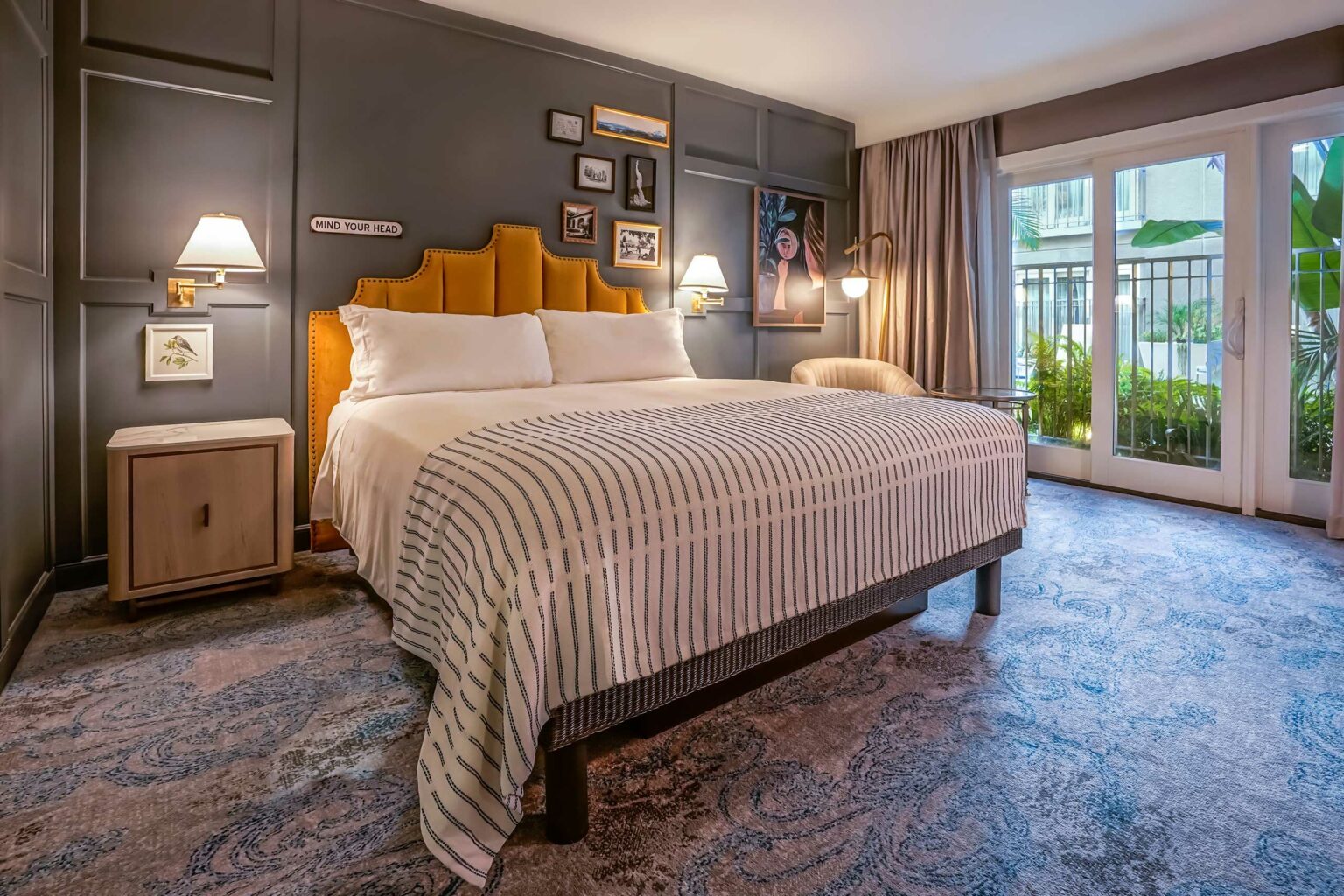 Hotel room with a cozy atmosphere, featuring a king-sized bed with a striped blanket, mustard headboard, framed art, soft lighting, and garden view.