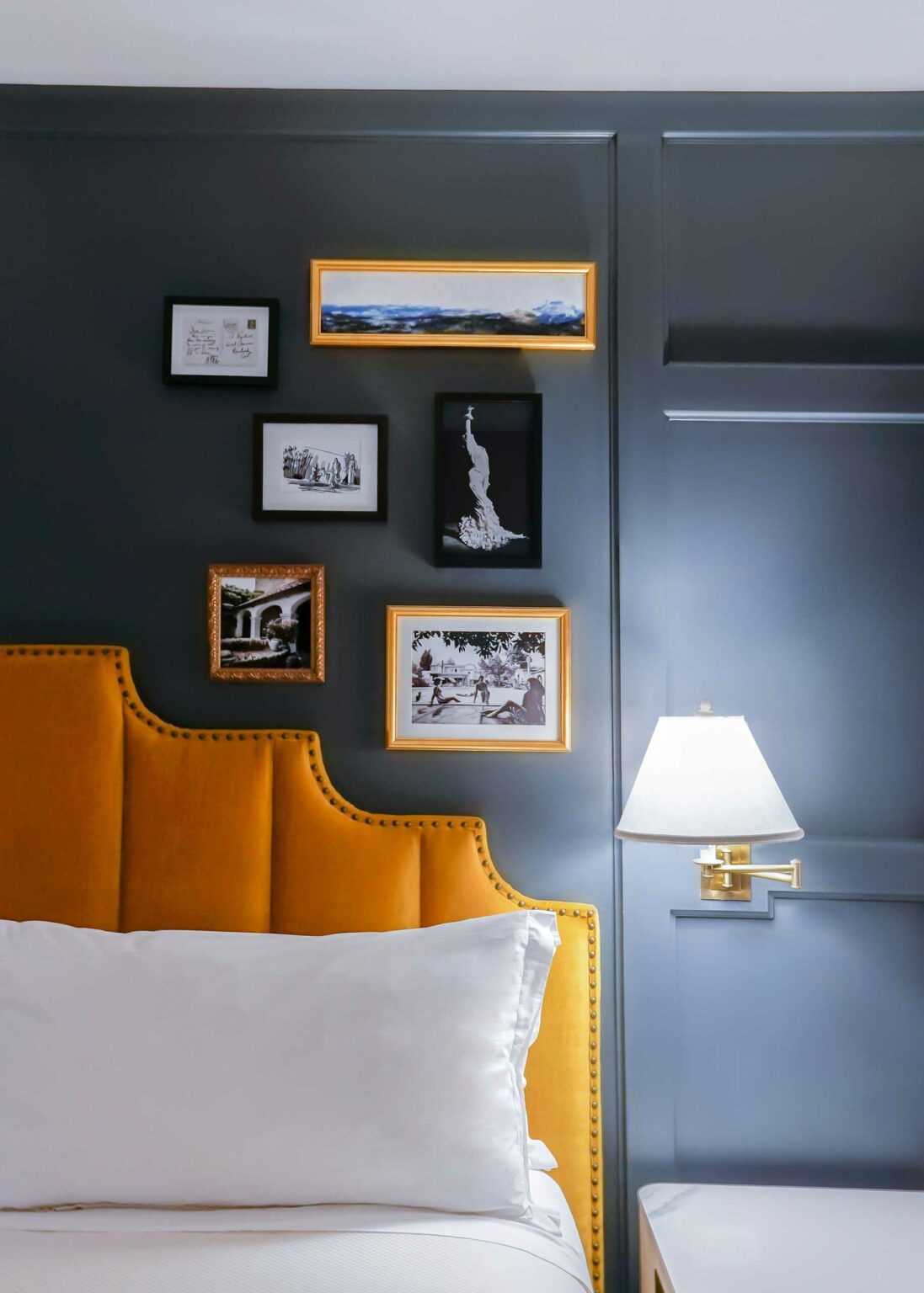 Stylish hotel bedroom wall with dark paneling, adorned with various art prints in gold and black frames above a mustard headboard and a lit wall lamp.